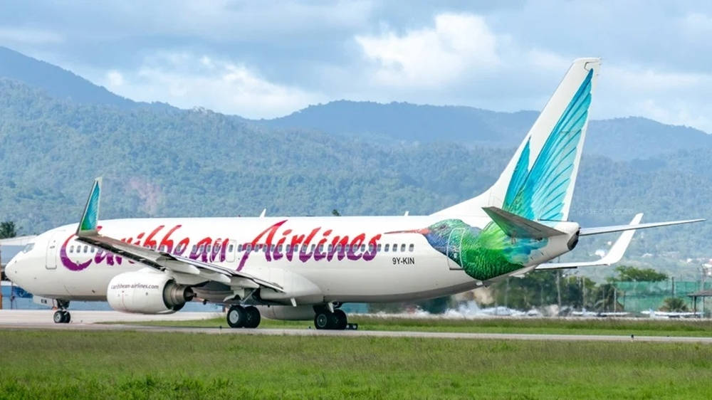  壁紙 — 一架加勒比航空的飛機帶有鮮豔的蜂鳥塗裝，在部分多雲的天空下，以鬱鬱蔥蔥的綠色山脈為背景，在跑道上滑行。
