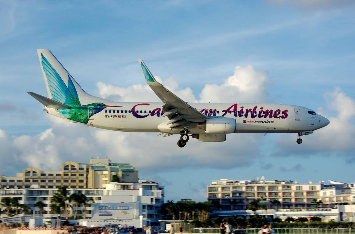 飛機 桌布 壁紙 — 一架加勒比航空的飛機放下起落架，在部分多雲的天空下低飛越過沿海酒店。