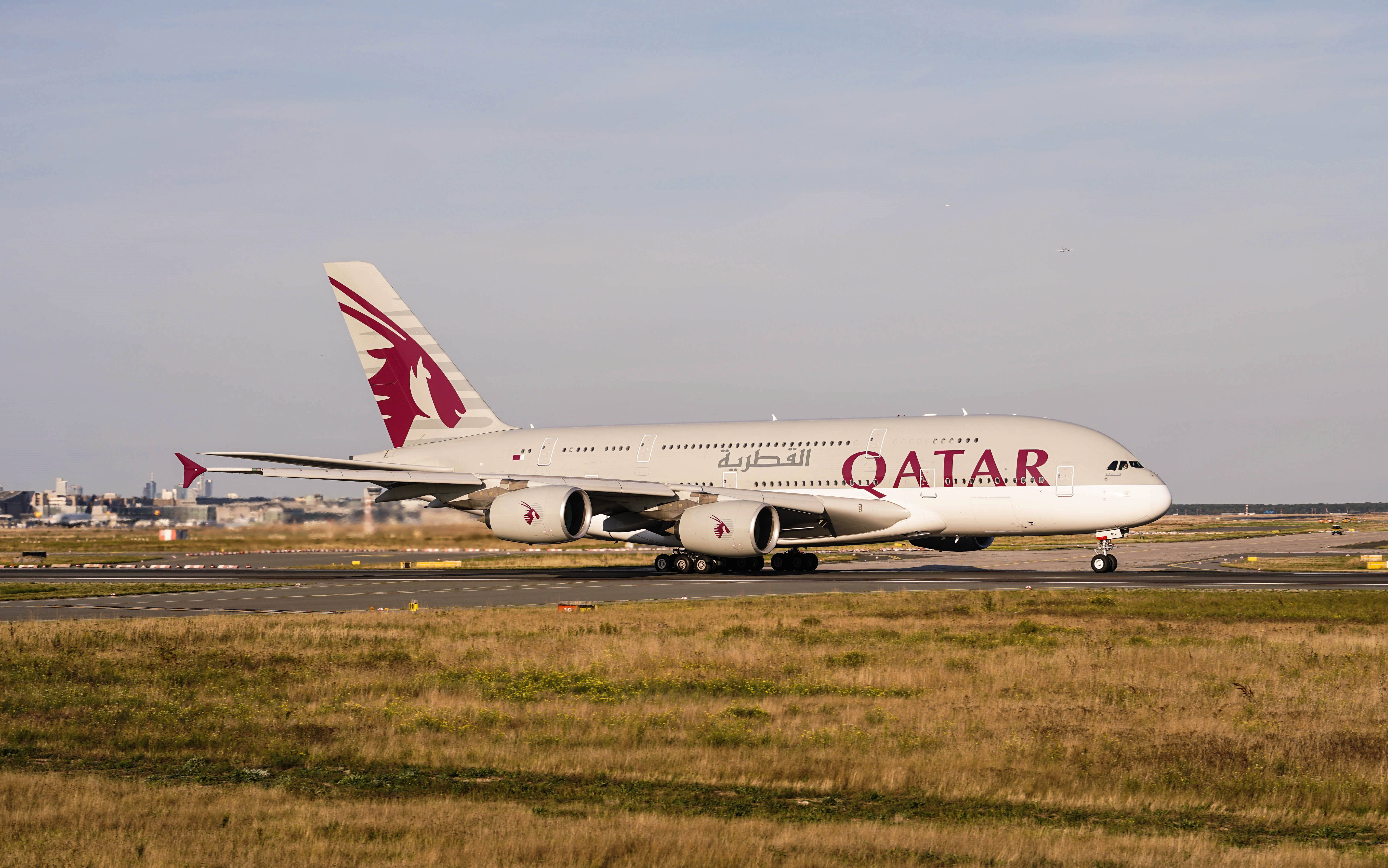 4K 飛機壁紙 桌布 壁紙 — 一架卡塔爾航空的空中巴士A380在乾草和遠處城市景觀的跑道上滑行，天空蒼白。