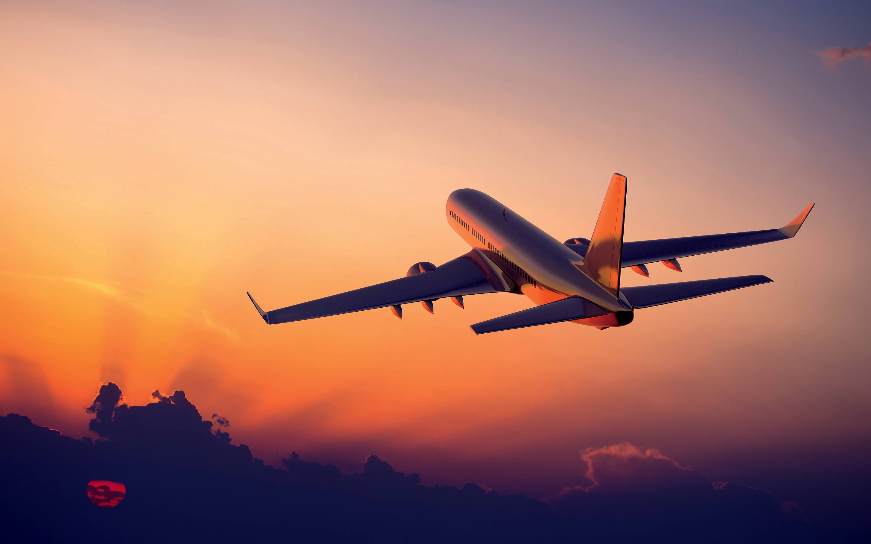 飛機 桌布 壁紙 — 一架商用飛機在充滿活力的日落天空中翱翔，天空呈現橙色和紫色色調，下方有輪廓分明的雲層。