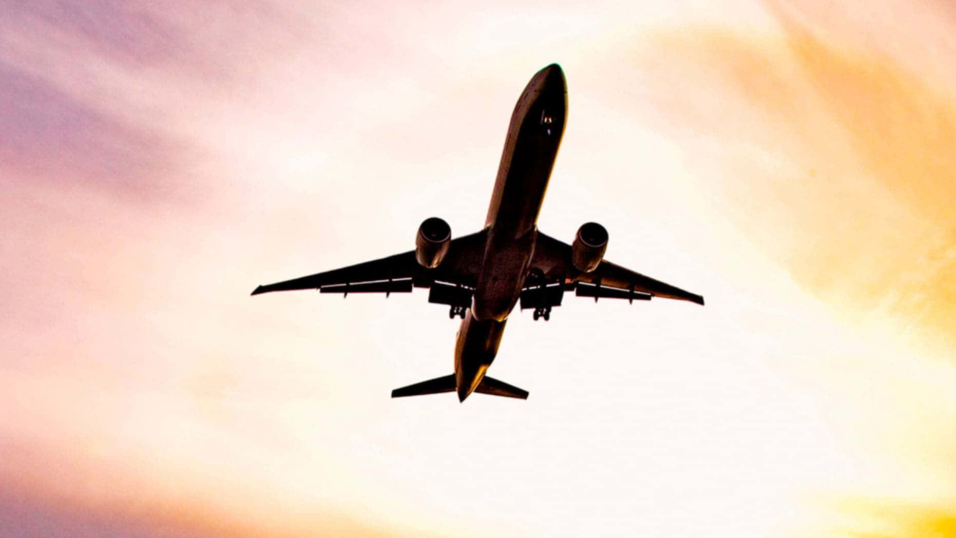 飛機 桌布 壁紙 — 一架商用飛機在充滿活力的日落天空襯托下形成剪影，天空中有橙色和粉紅色的雲彩條紋。