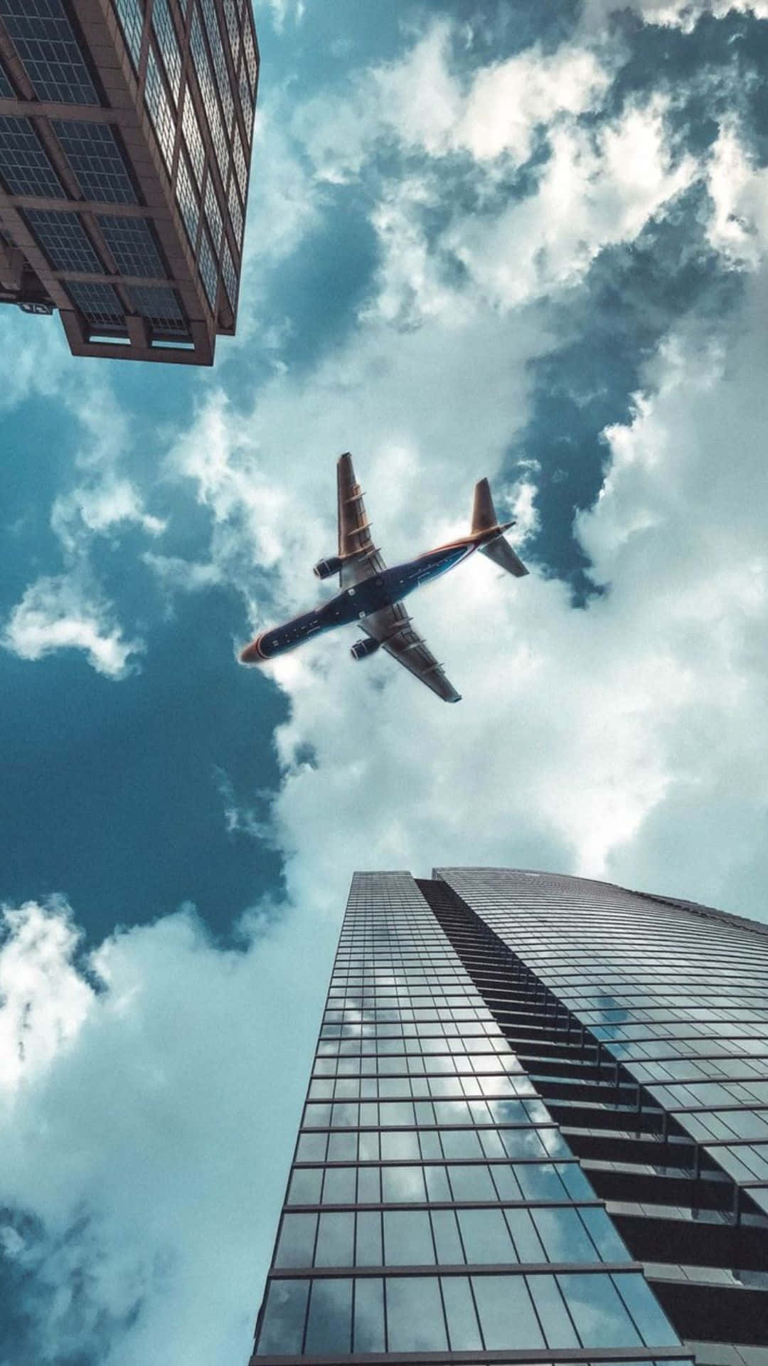 Android Small Planes 桌布 壁紙 — 一架商用飛機在戲劇性的多雲天空下，翱翔於高聳的玻璃摩天大樓之間。