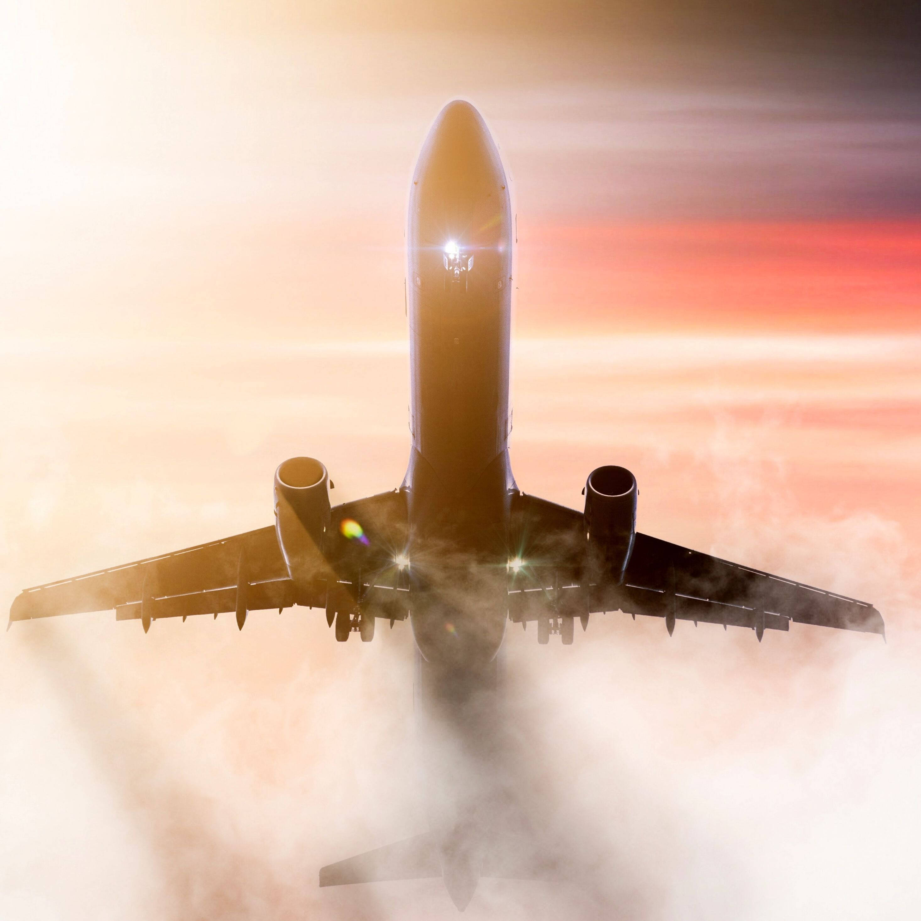 日落 桌布 壁紙 — 一架商用飛機在日出時翱翔於雲層之上，引擎發光，天空呈現火熱的橙色。