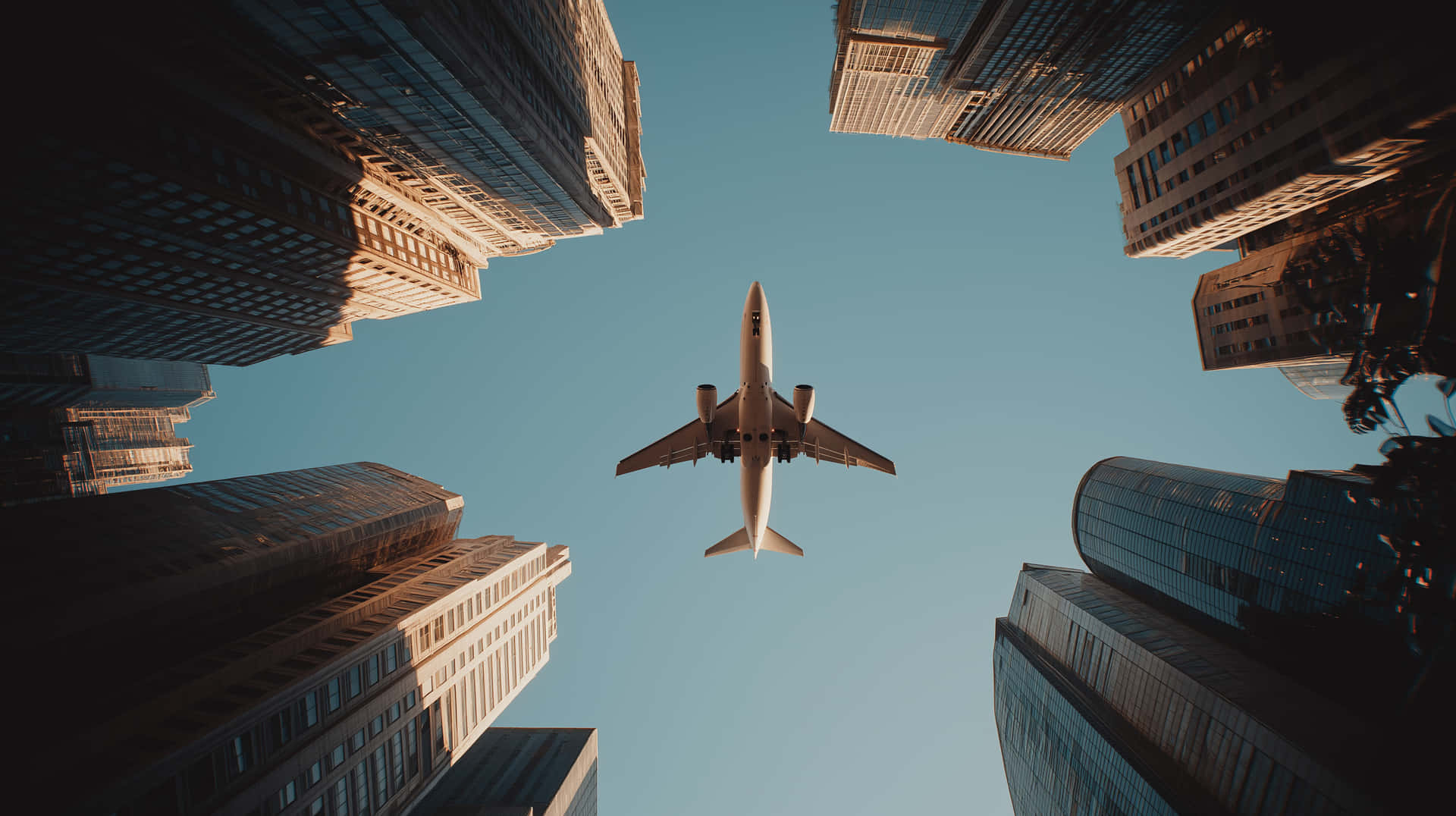 Cinematic View of a Plane Soaring Between Towering City Skyscrapers 壁紙 — 一架商用飛機在晴朗的藍天下翱翔，周圍是高聳的玻璃和鋼鐵摩天大樓。