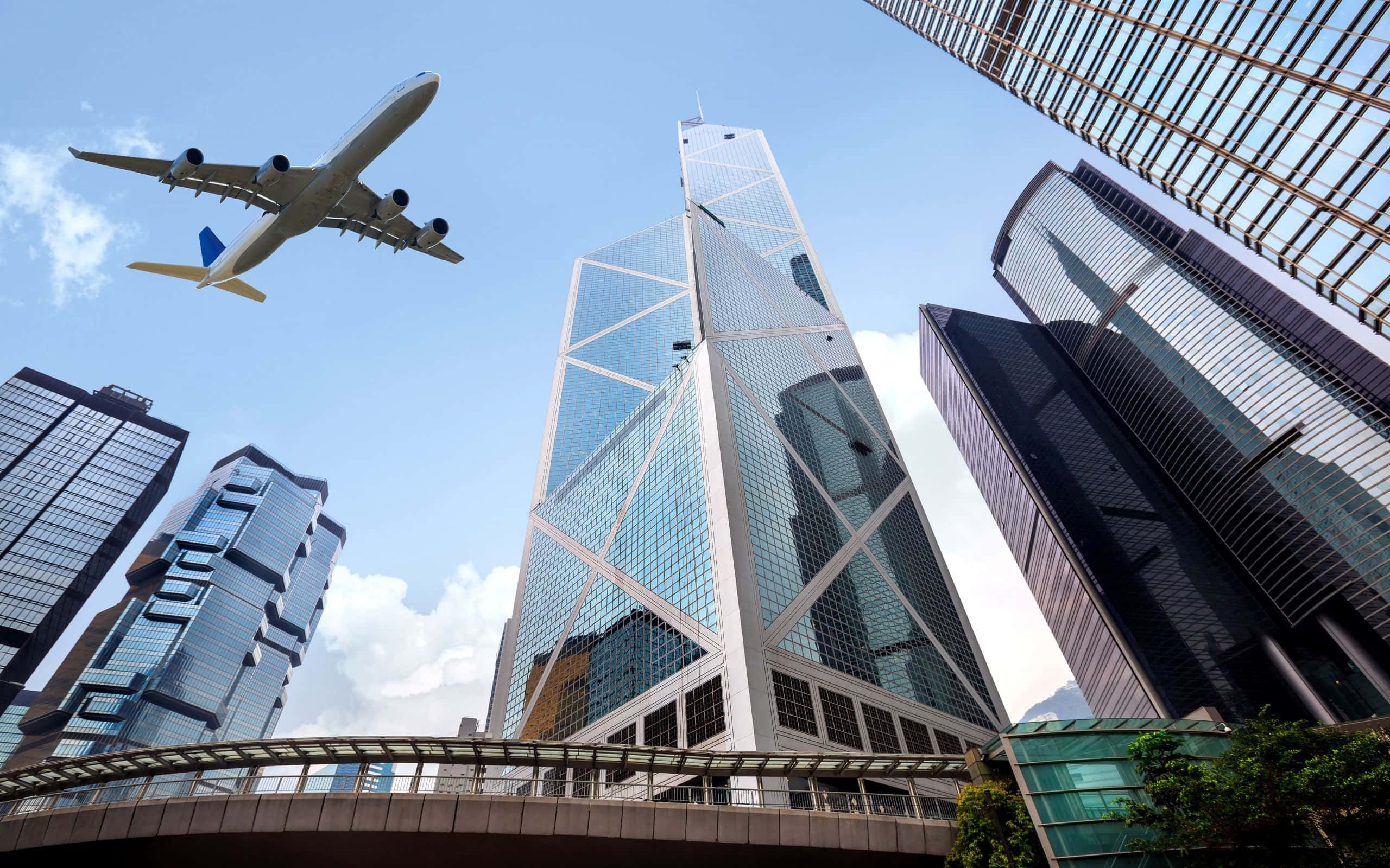 Bank 桌布 壁紙 — 一架商用飛機在晴朗藍天與零星白雲下，翱翔於高聳的玻璃摩天大樓之上。