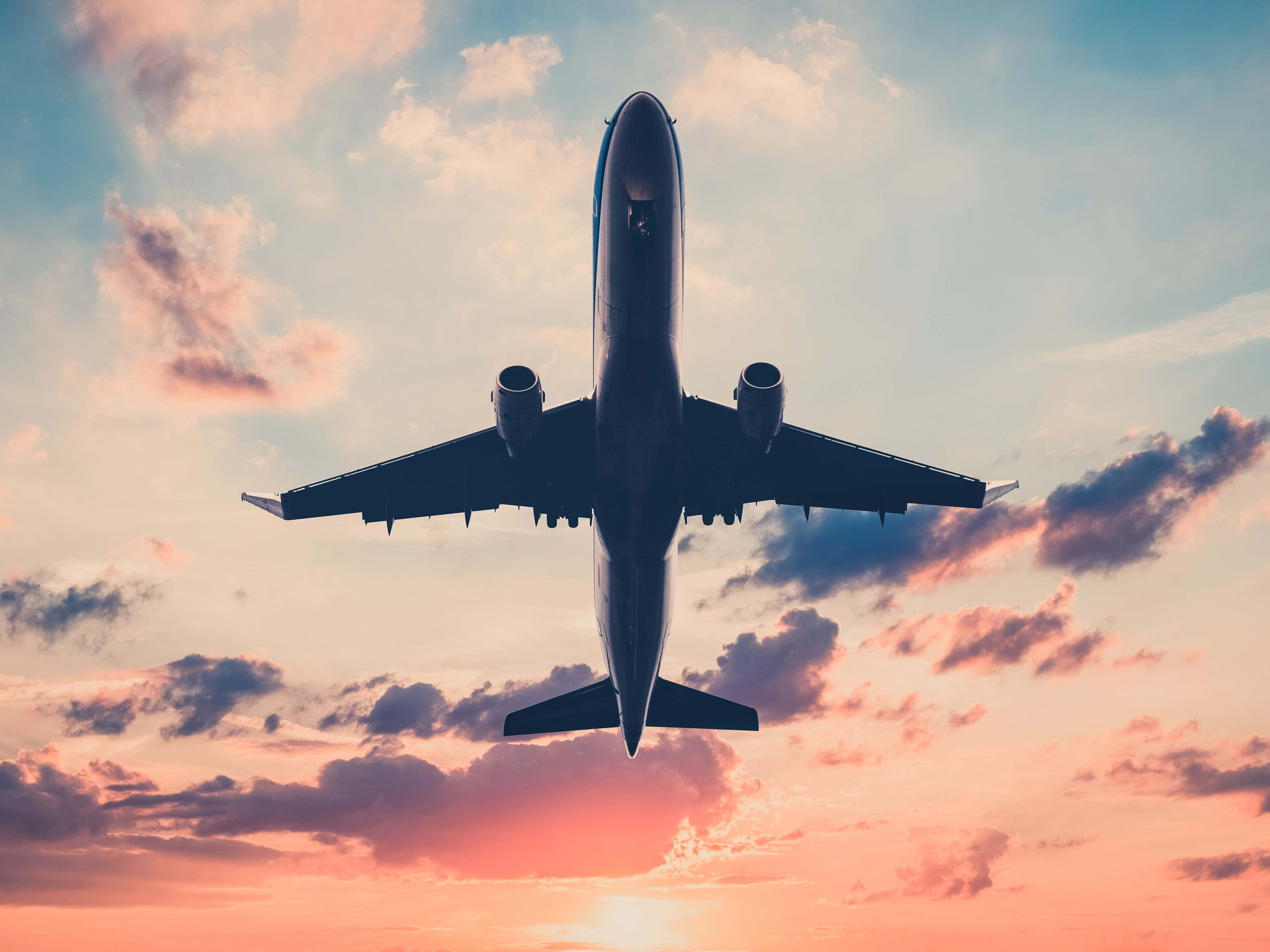 Travel Desktop 桌布 壁紙 — 一架商用飛機在頭頂翱翔，背景是充滿活力的日落天空，塗有粉紅色、橙色和藍色的雲彩。