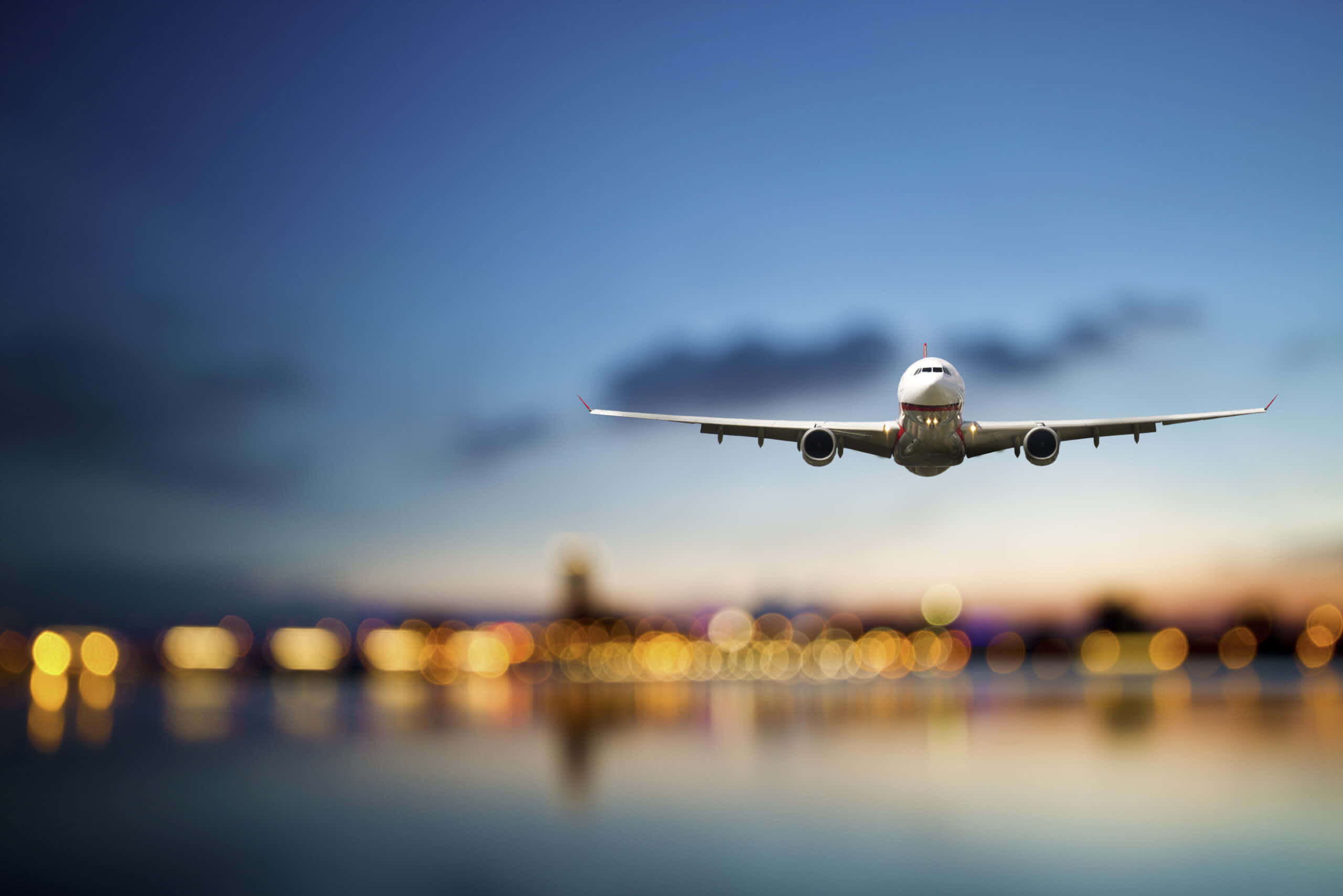 Travel Desktop 桌布 壁紙 — 一架商用飛機在黃昏時低飛過水面，下方模糊的城市燈光倒映著。