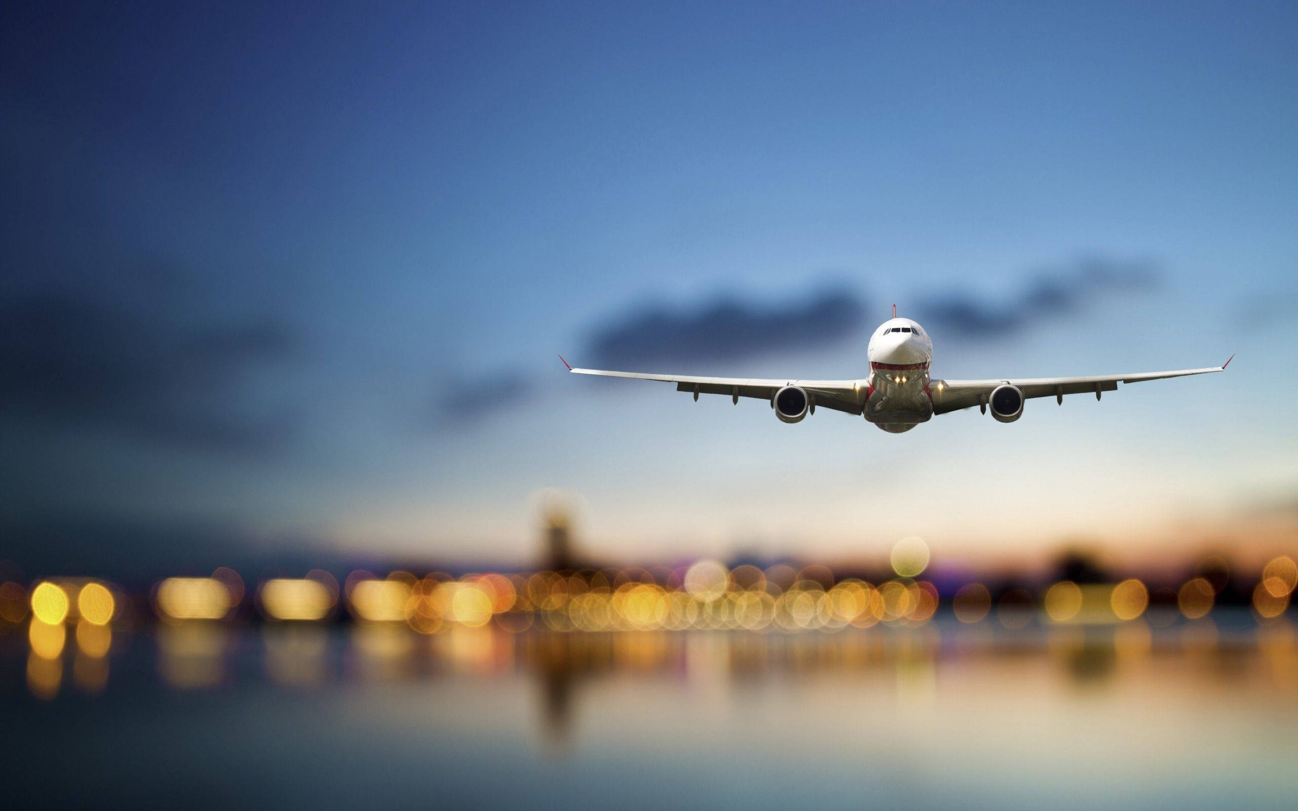 4K 飛機壁紙 桌布 壁紙 — 一架商用飛機在黃昏時分向城市天際線下降，模糊的金色燈光倒映在下方水面上。