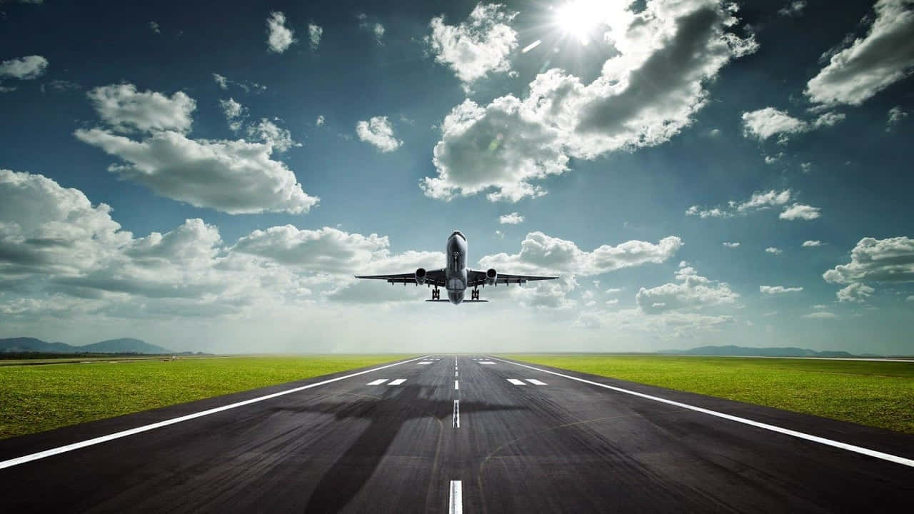 飛機 桌布 壁紙 — 一架商用飛機從跑道升起，跑道兩旁是充滿活力的綠色田野，上方是明亮的陽光和散落的雲朵。