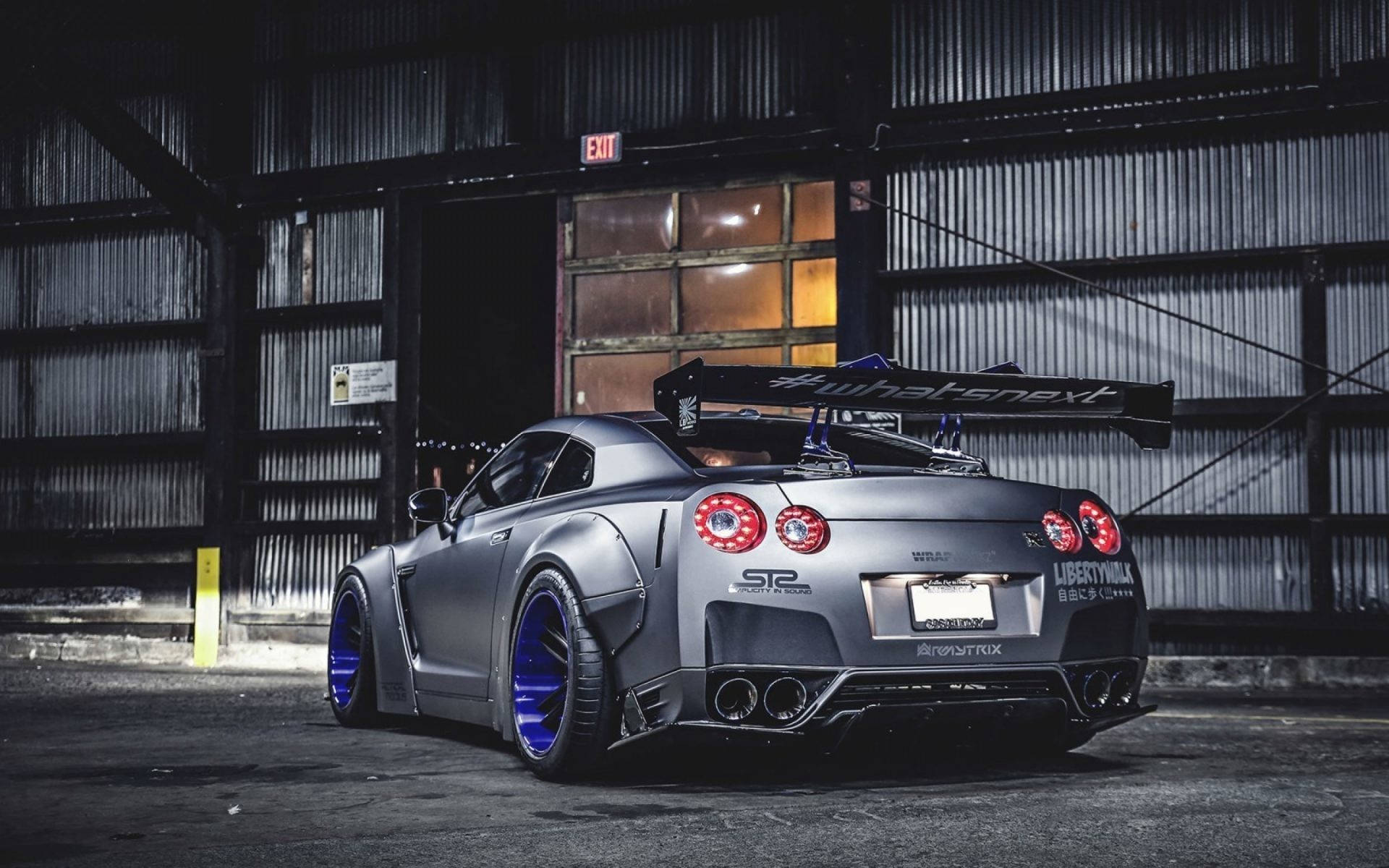 Nissan 桌布 壁紙 — 一架啞光灰色日產GT-R，配備藍色輪圈和大型尾翼，停放在燈光昏暗的工業倉庫內。