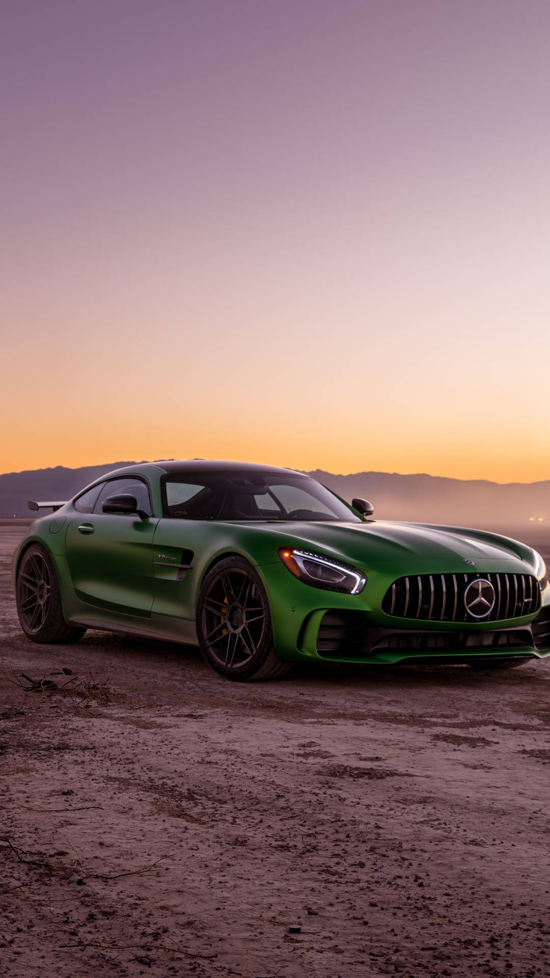 賓士 iPhone 桌布 壁紙 — 一架啞光綠色的Mercedes-AMG GT停泊在塵土飛揚的地形上，背景有山脈和日落。
