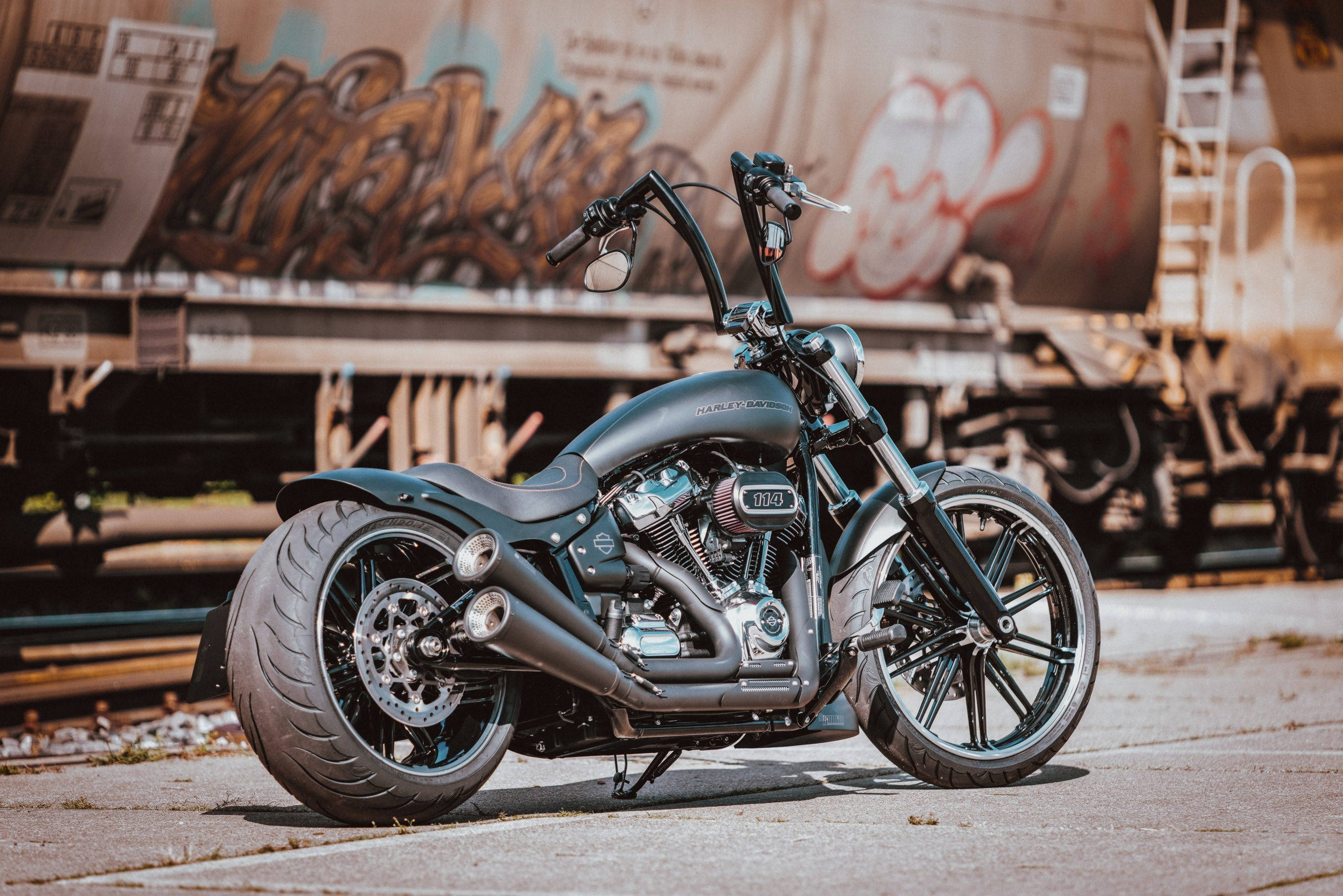 Chopper 桌布 壁紙 — 一架啞黑色訂製chopper機車停在混凝土上，旁邊是塗滿塗鴉的火車車廂，在溫暖的陽光下。