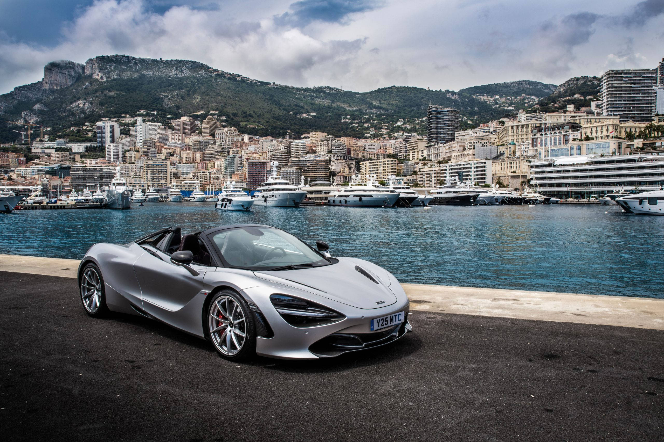 跑車 桌布 壁紙 — 一架型格銀色McLaren 720S Spider停泊在海濱長廊，背景是豪華遊艇和密集的沿海城市景觀。
