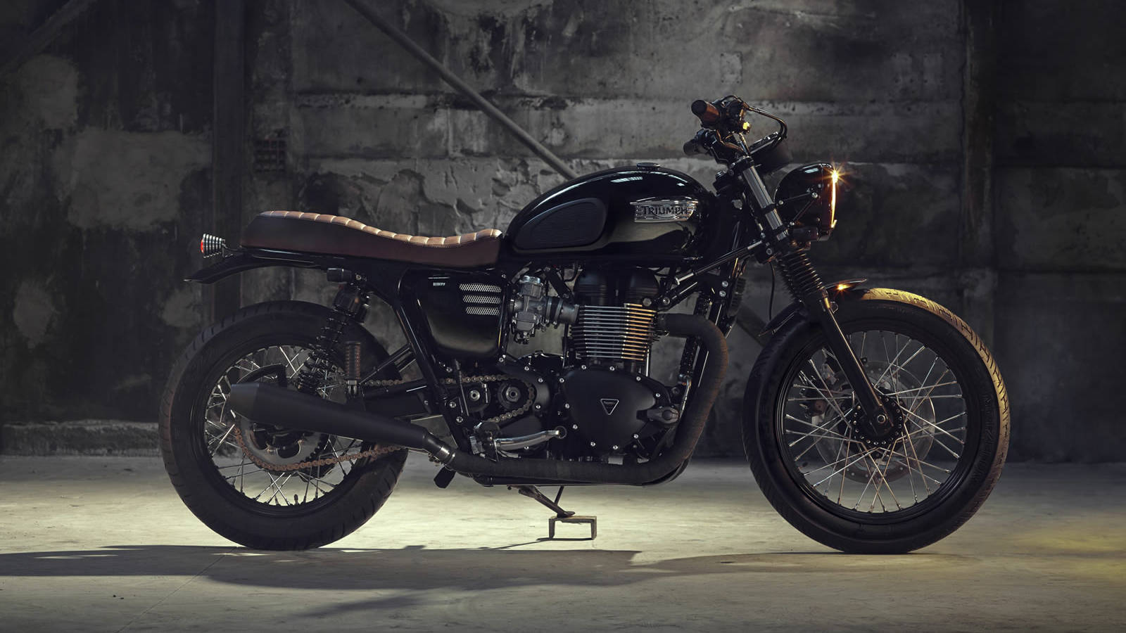 Triumph 桌布 壁紙 — 一架型格黑色Triumph機車搭配棕色皮革座椅，在燈光昏暗的混凝土車庫中，被聚光燈戲劇性地照亮。