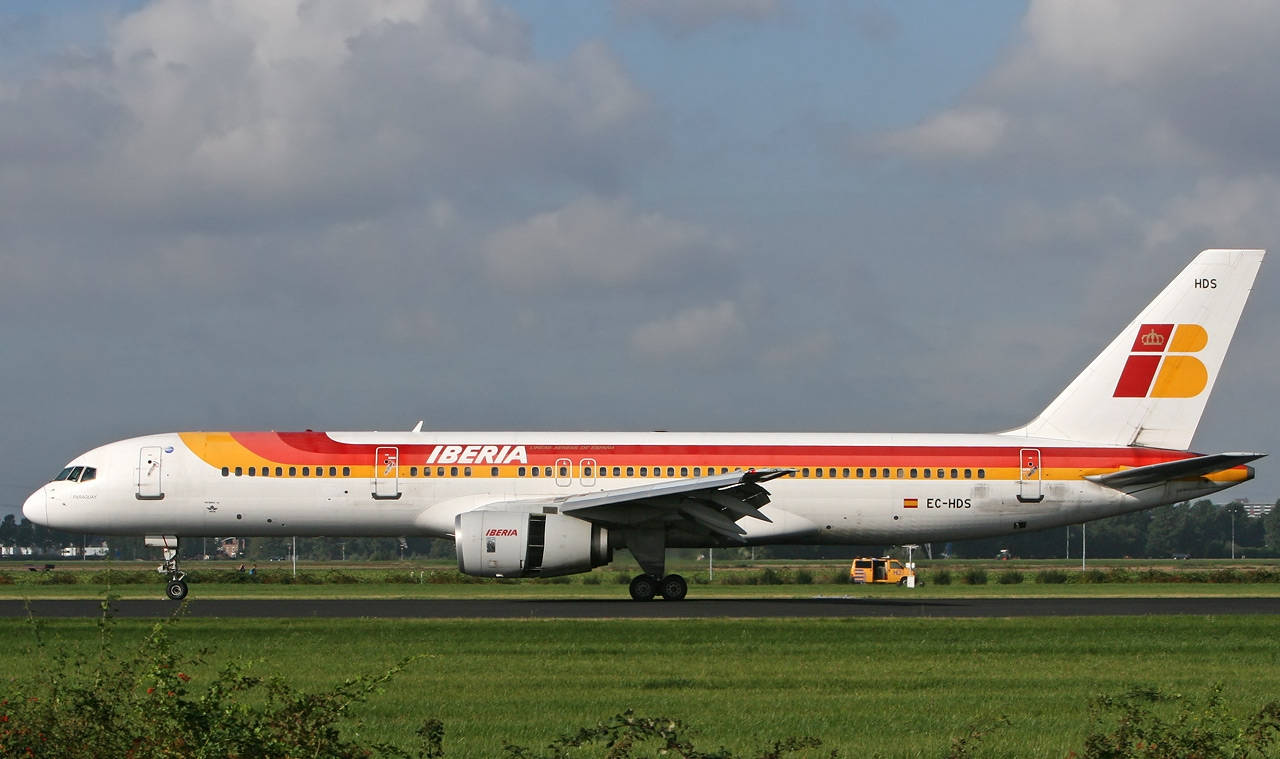 飛機 桌布 壁紙 — 一架塗有紅、黃、白色塗裝的伊比利亞航空飛機，在陰天、綠草環繞的跑道上滑行。