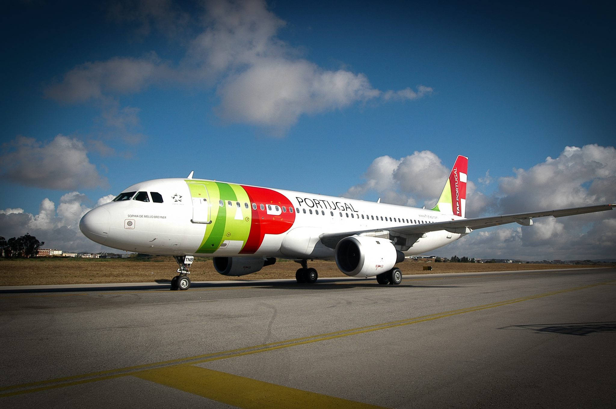 Airport 桌布 壁紙 — 一架塗有鮮豔綠色、紅色和白色塗裝的TAP Portugal飛機，在部分多雲的藍天下，於陽光明媚的機場停機坪上滑行。