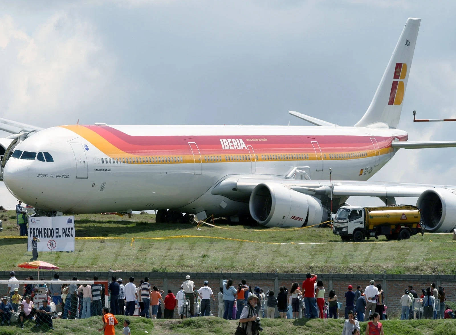 飛機 桌布 壁紙 — 一架大型伊比利亞航空空中巴士A340飛機停泊在草地上，附近有大批圍觀人群和一輛油車，天空多雲。