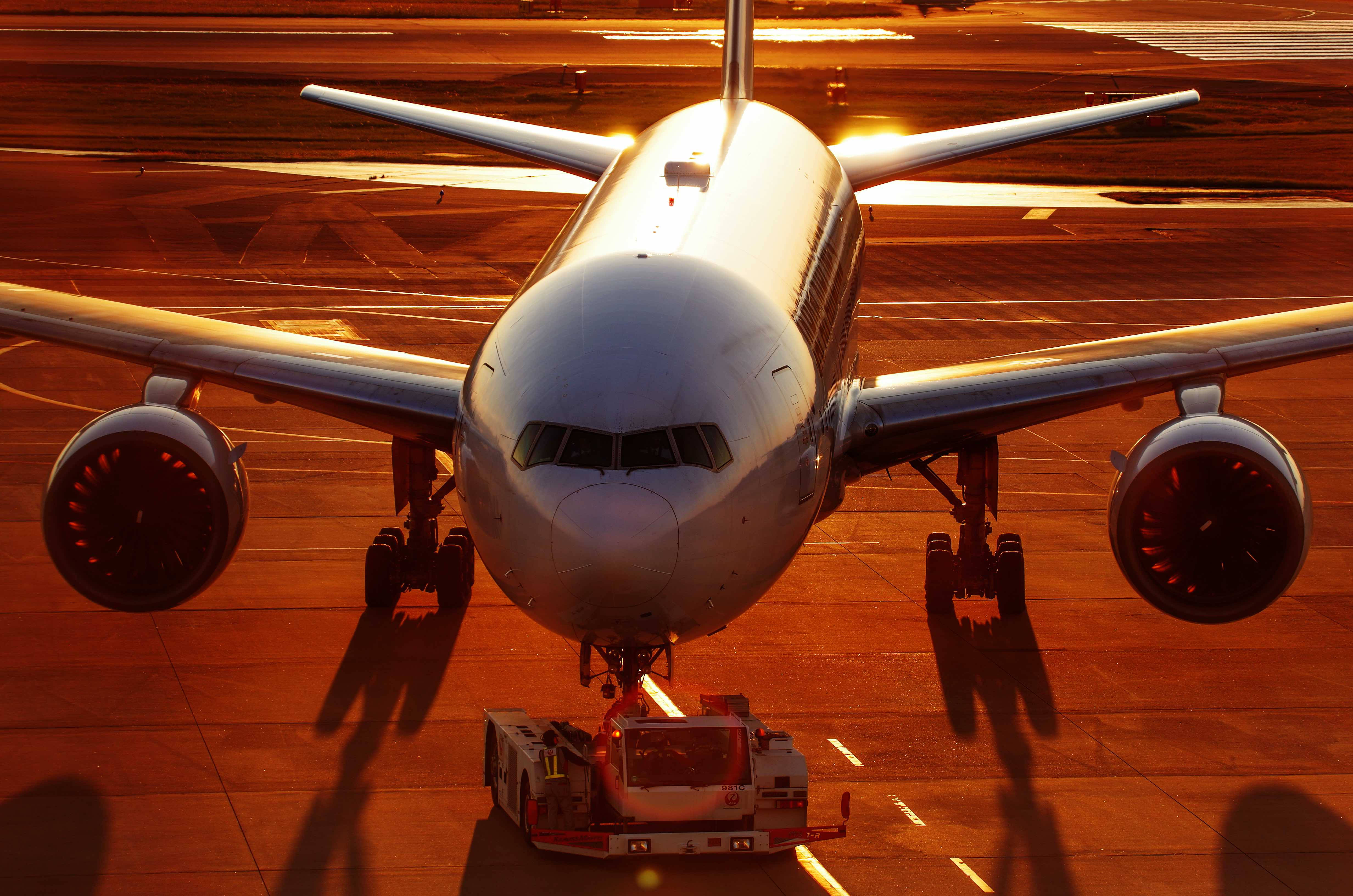 飛機 桌布 壁紙 — 一架大型客機停在機場停機坪上，沐浴在金色的日落光芒中，機頭位置有一輛牽引車。