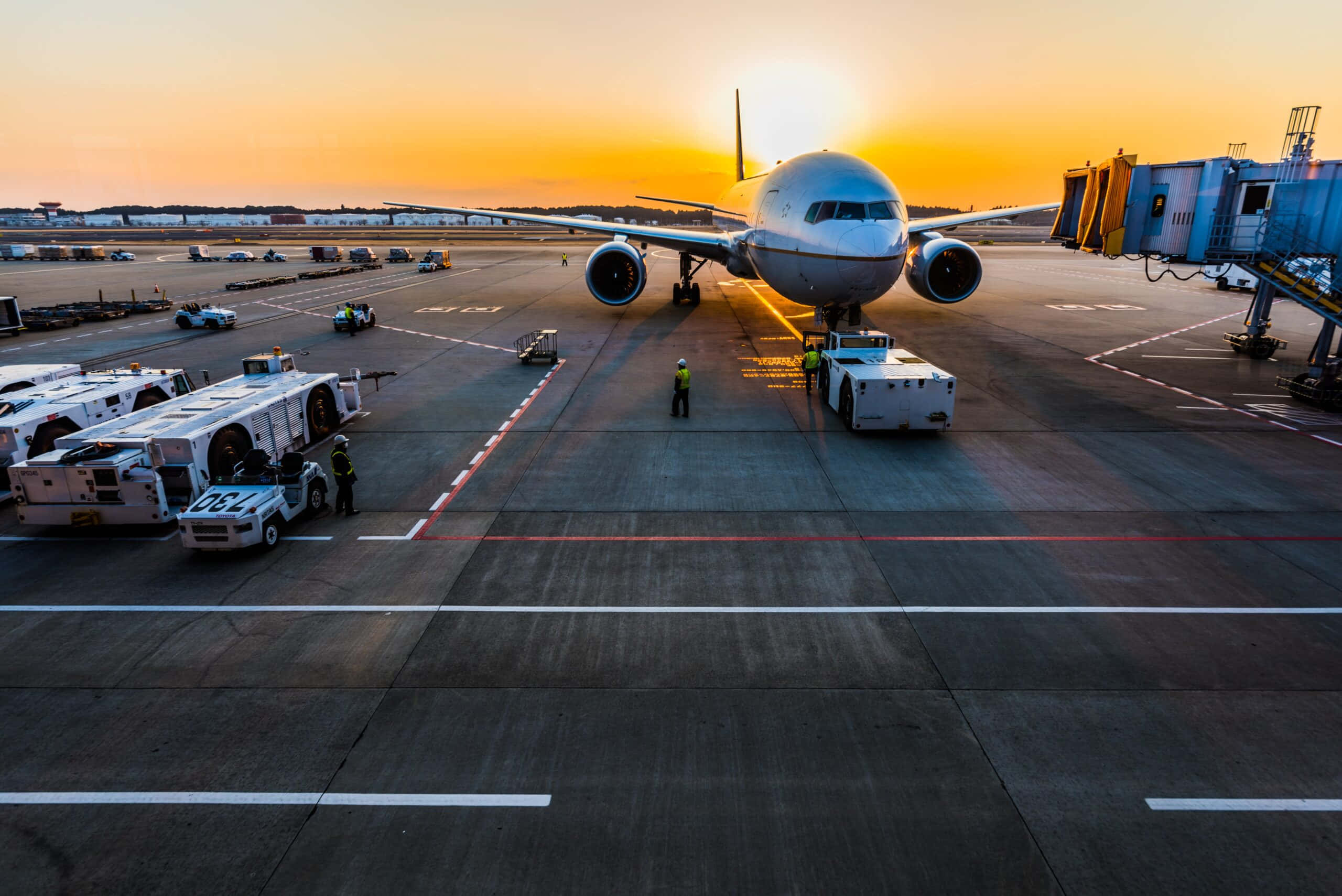 Airport 桌布 壁紙 — 一架大型客機在充滿活力的橙色日落時分停在機場閘口，附近有地勤人員和服務車輛。