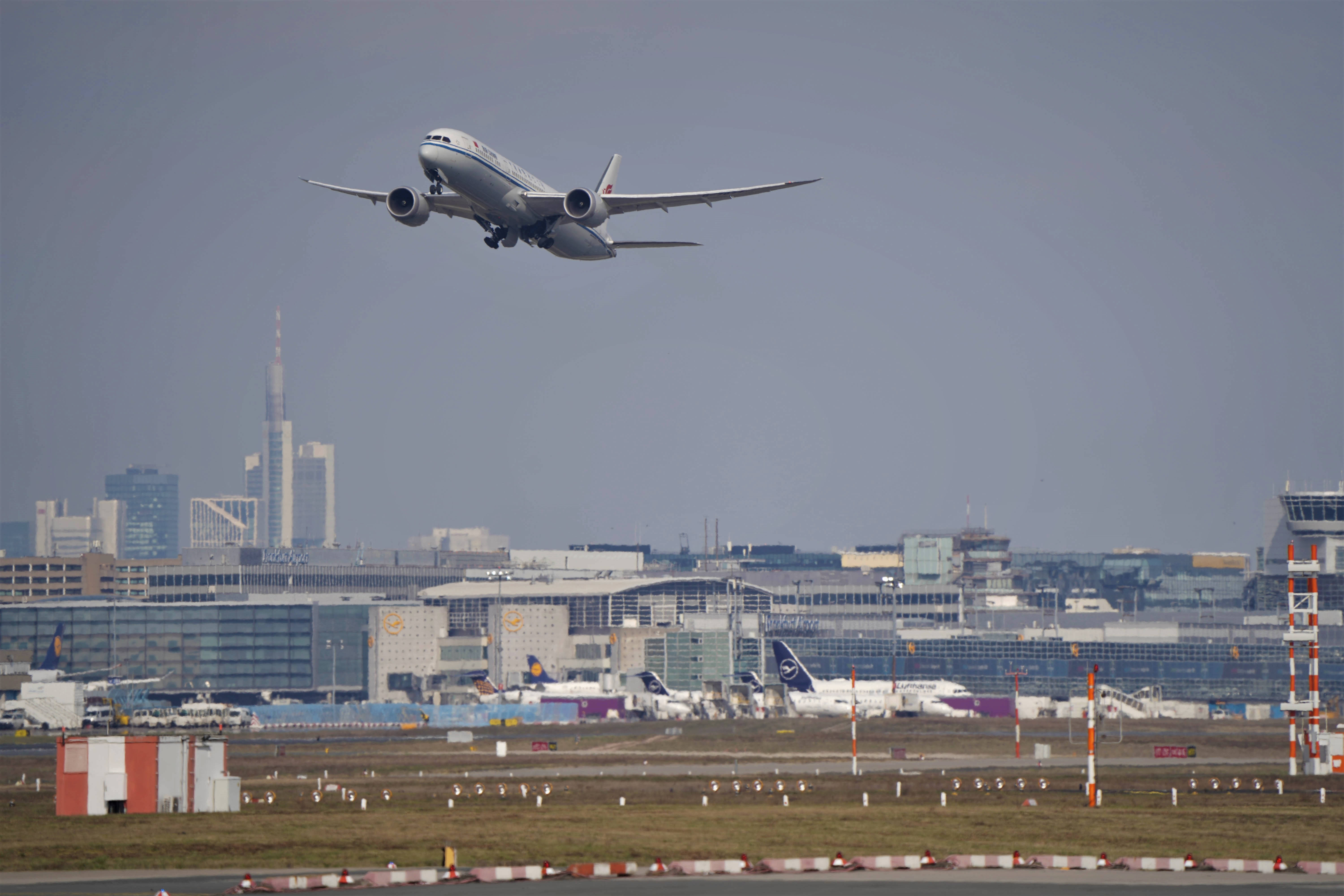 4K 飛機壁紙 桌布 壁紙 — 一架大型客機在廣闊的現代化機場航廈上空，攀升至煙霧瀰漫的天空，背景是城市天際線。