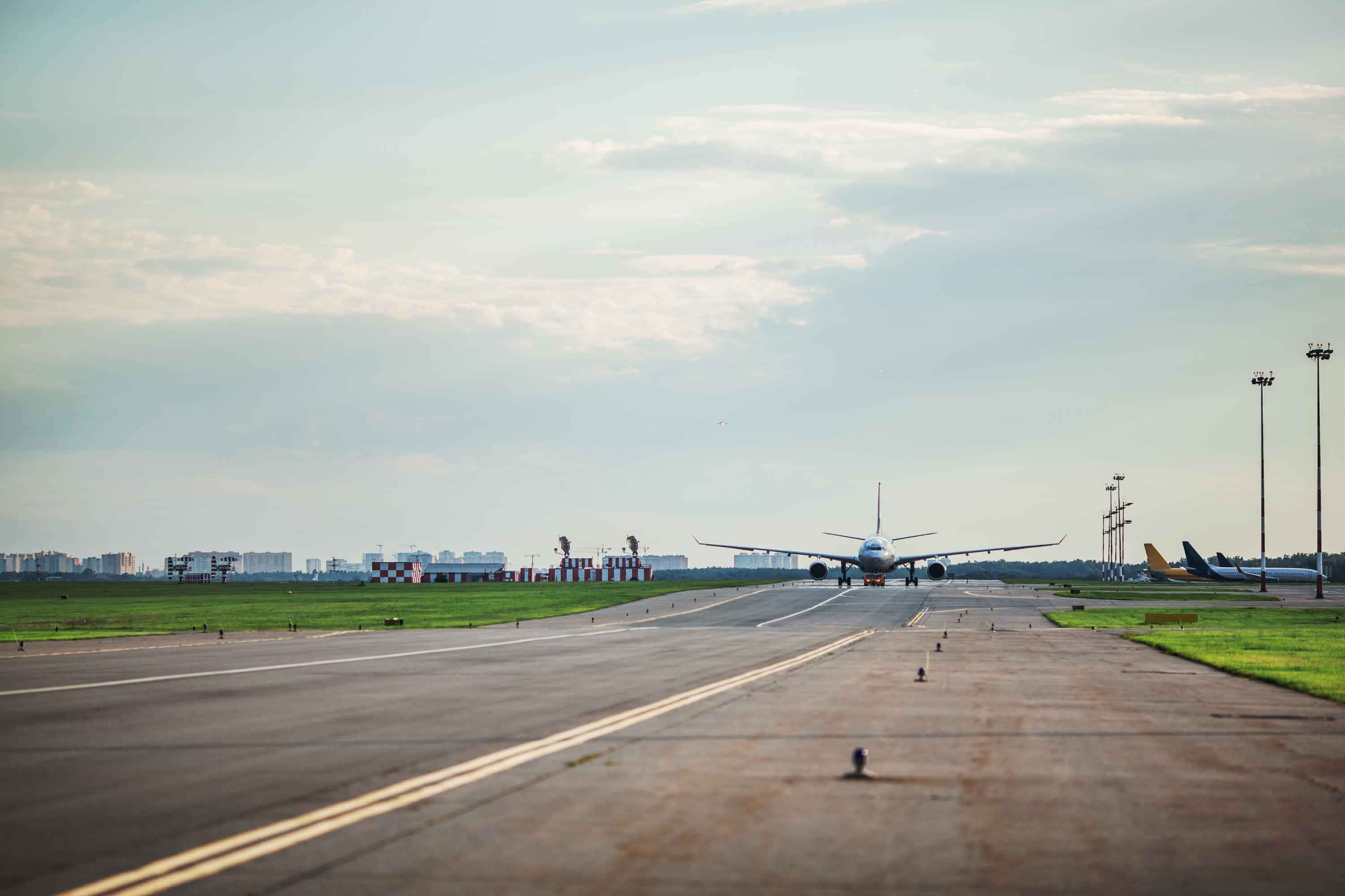 Runway 桌布 壁紙 — 一架大型客機在柔和的多雲天空下，沿著長長的機場跑道滑行，遠處有城市建築。