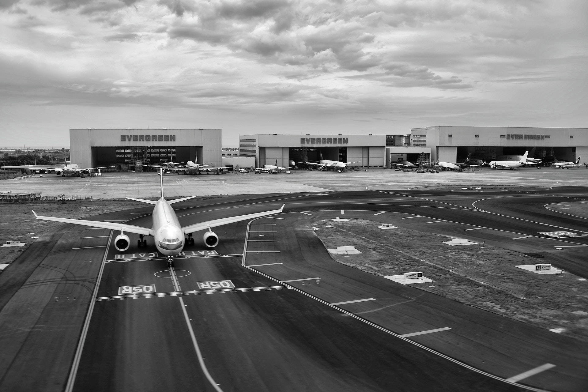 Runway 桌布 壁紙 — 一架大型客機在機場停機坪上的黑白航拍圖，背景有機庫和其他飛機，天空多雲。