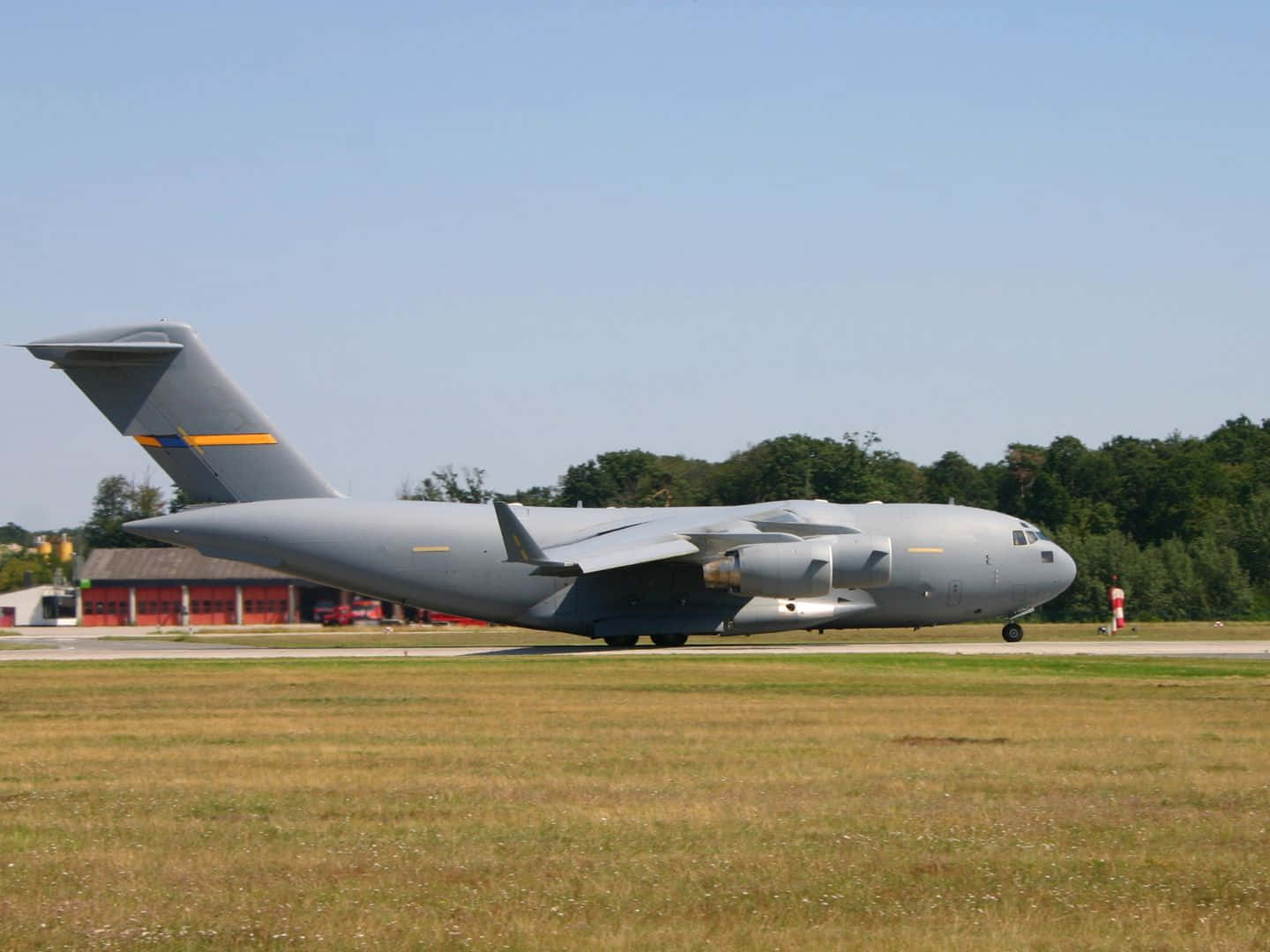 Military Plane 桌布 壁紙 — 一架大型灰色軍用運輸機在晴朗藍天下，以綠色樹木和紅色機庫為背景，在跑道上滑行。
