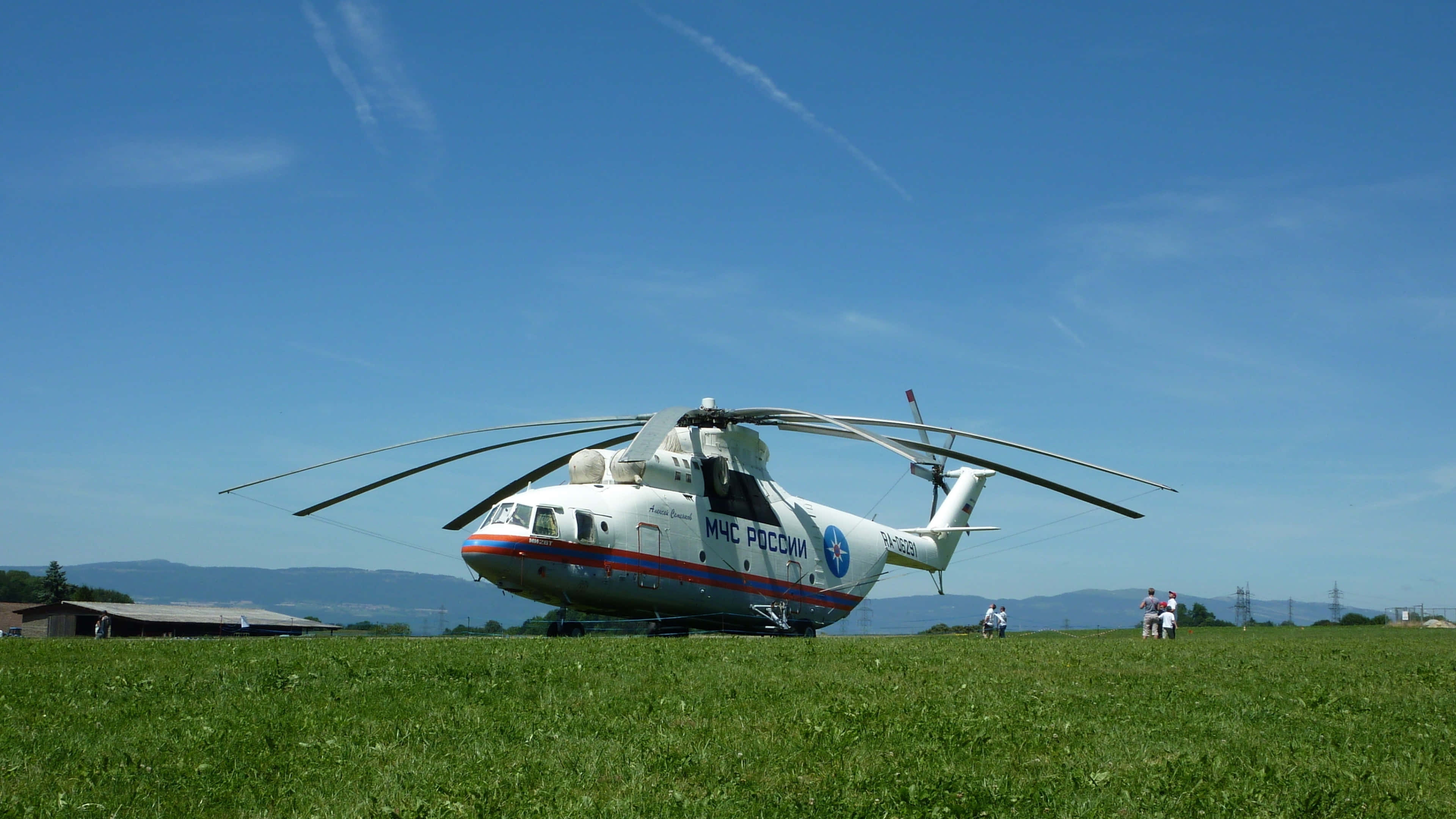 4K Helicopters 桌布 壁紙 — 一架大型白色俄羅斯救援直升機停放在一片充滿活力的綠色田野上，背景是晴朗的藍天和遠處的山脈。