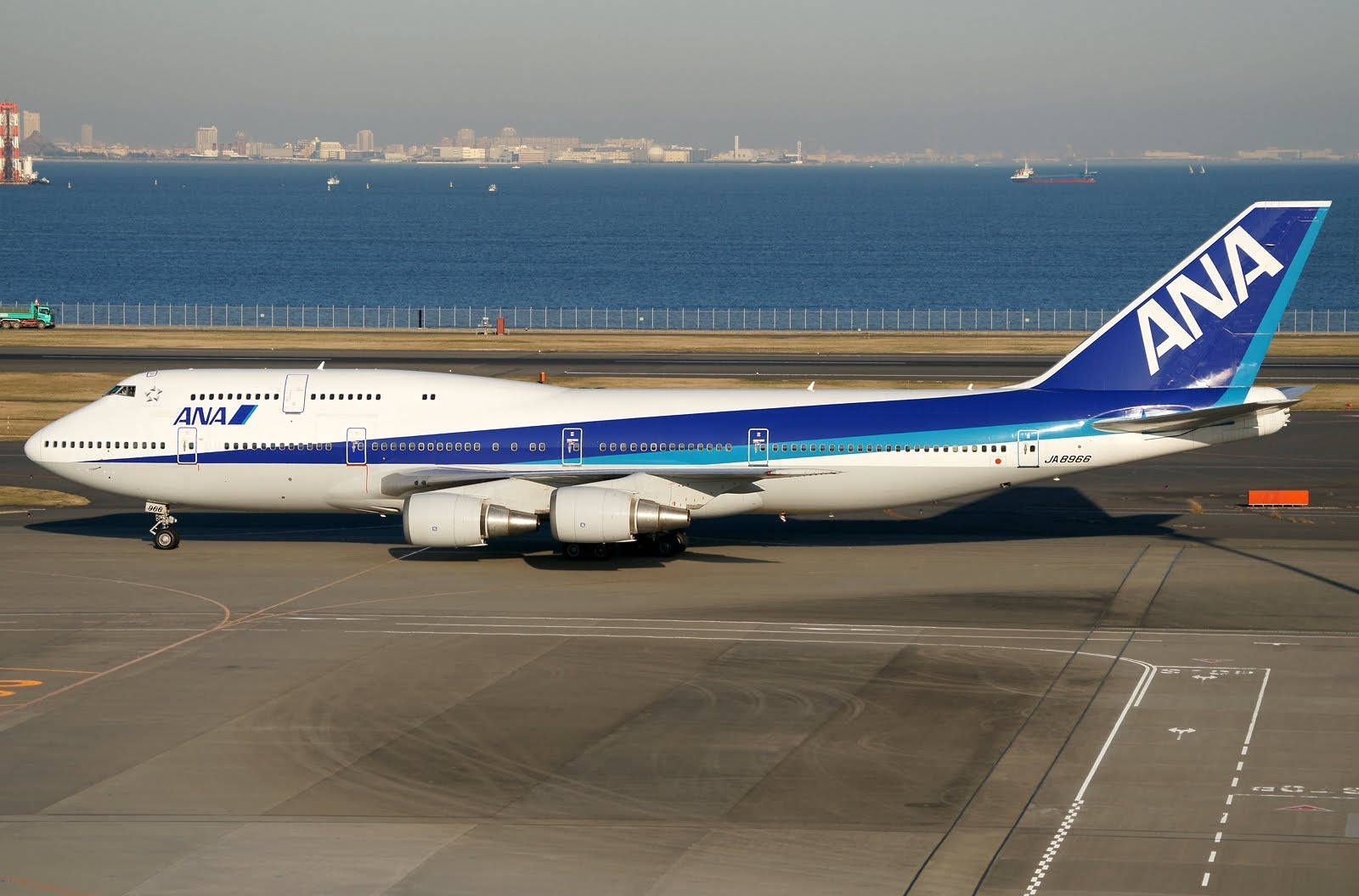 Airport 桌布 壁紙 — 一架大型白色和藍色的ANA波音747停放在跑道上，旁邊是廣闊的藍色海洋，遠處的城市天際線在朦朧的天空下。