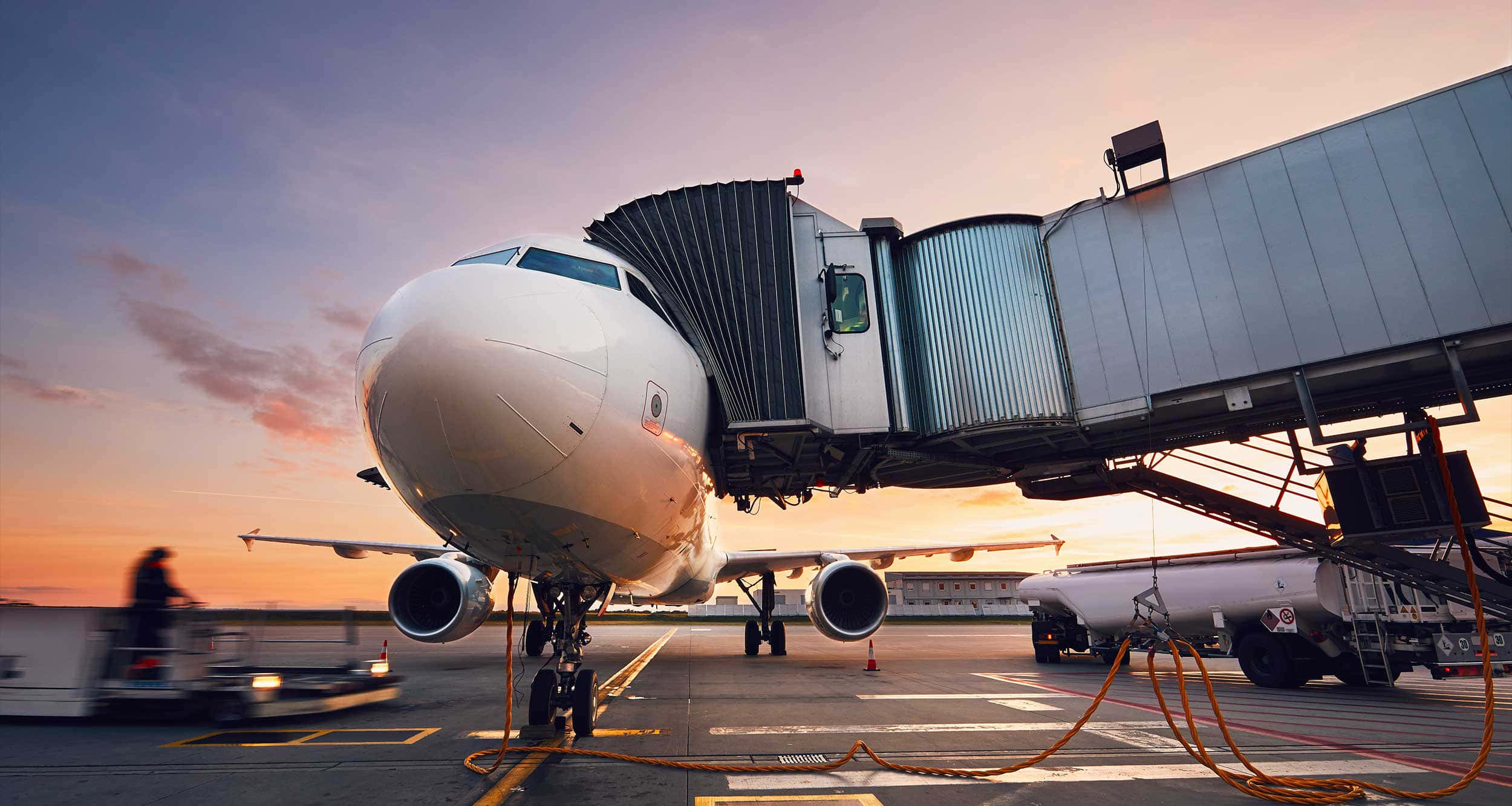飛機 桌布 壁紙 — 一架大型白色客機停在機場閘口，連接了登機橋，夕陽以溫暖的橙色和粉紅色調落下。