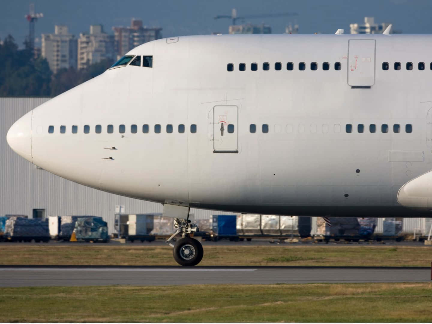 飛機 桌布 壁紙 — 一架大型白色客機停在跑道上，背景有城市建築物和貨櫃。