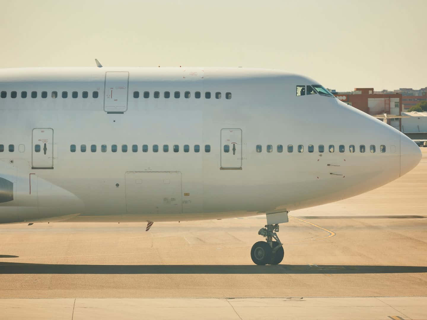 飛機 桌布 壁紙 — 一架大型白色客機靜止在陽光普照的機場停機坪上，背景有朦朧的建築物。