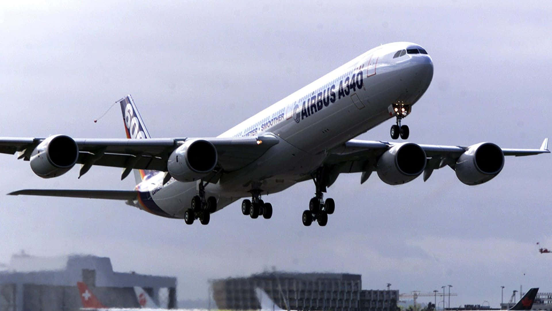 1920x1080 Jumbo Jets 桌布 壁紙 — 一架大型白色空中巴士A340飛機放下起落架，在陰天背景下接近降落，背景有城市建築物。