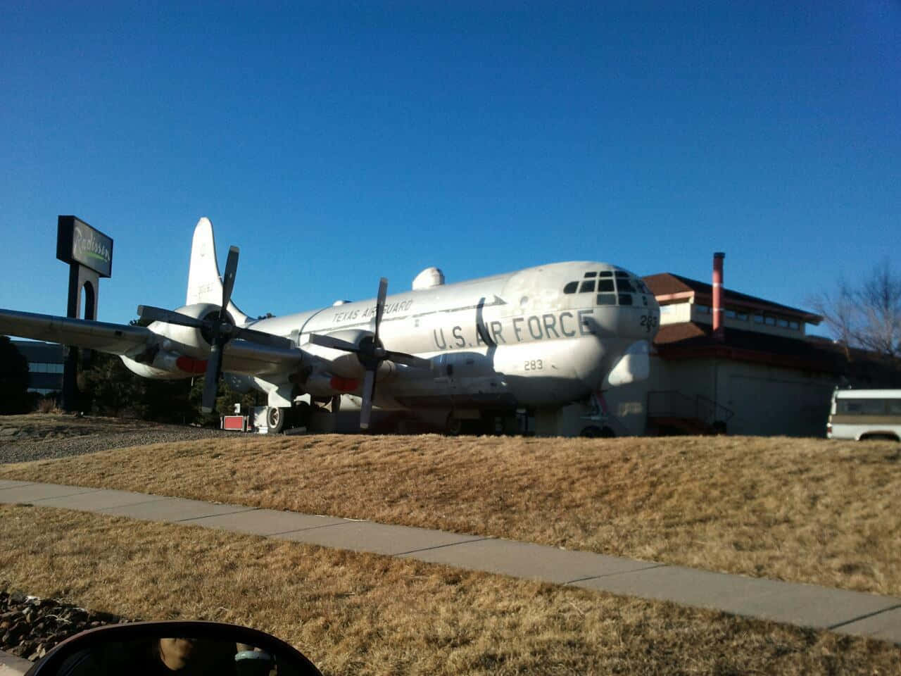 飛機 桌布 壁紙 — 一架大型白色美國空軍螺旋槳飛機停在乾草地上，靠近一棟建築物，天空清澈湛藍。