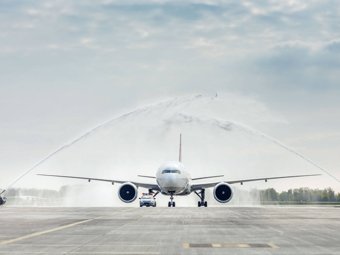 Airport 桌布 壁紙 — 一架大型白色飛機在灰色停機坪上，於多雲的天空下接受慶祝性的水砲致敬。