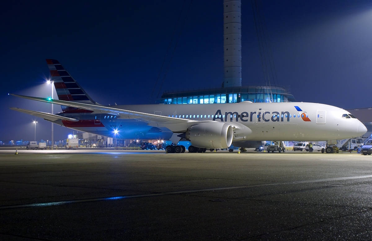 Airlines 桌布 壁紙 — 一架大型美國航空噴射機在夜間濕滑的停機坪上，在明亮的航廈燈光下閃閃發光，背景中控制塔聳立。