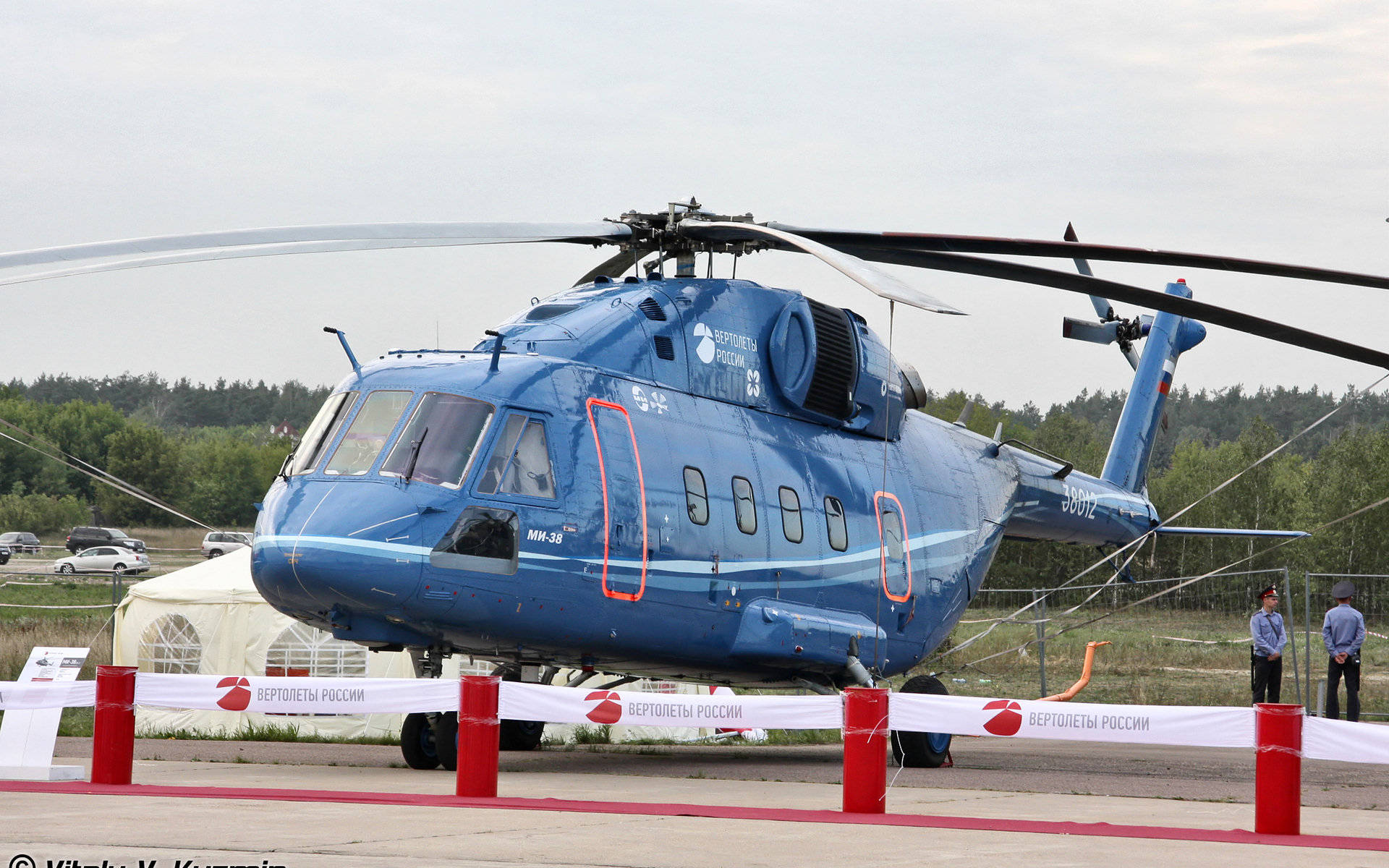 helicopter 4K 桌布 壁紙 — 一架大型藍色Mi-38直升機停在機場上，設有紅色障礙物，兩名男子在陰天附近站立。