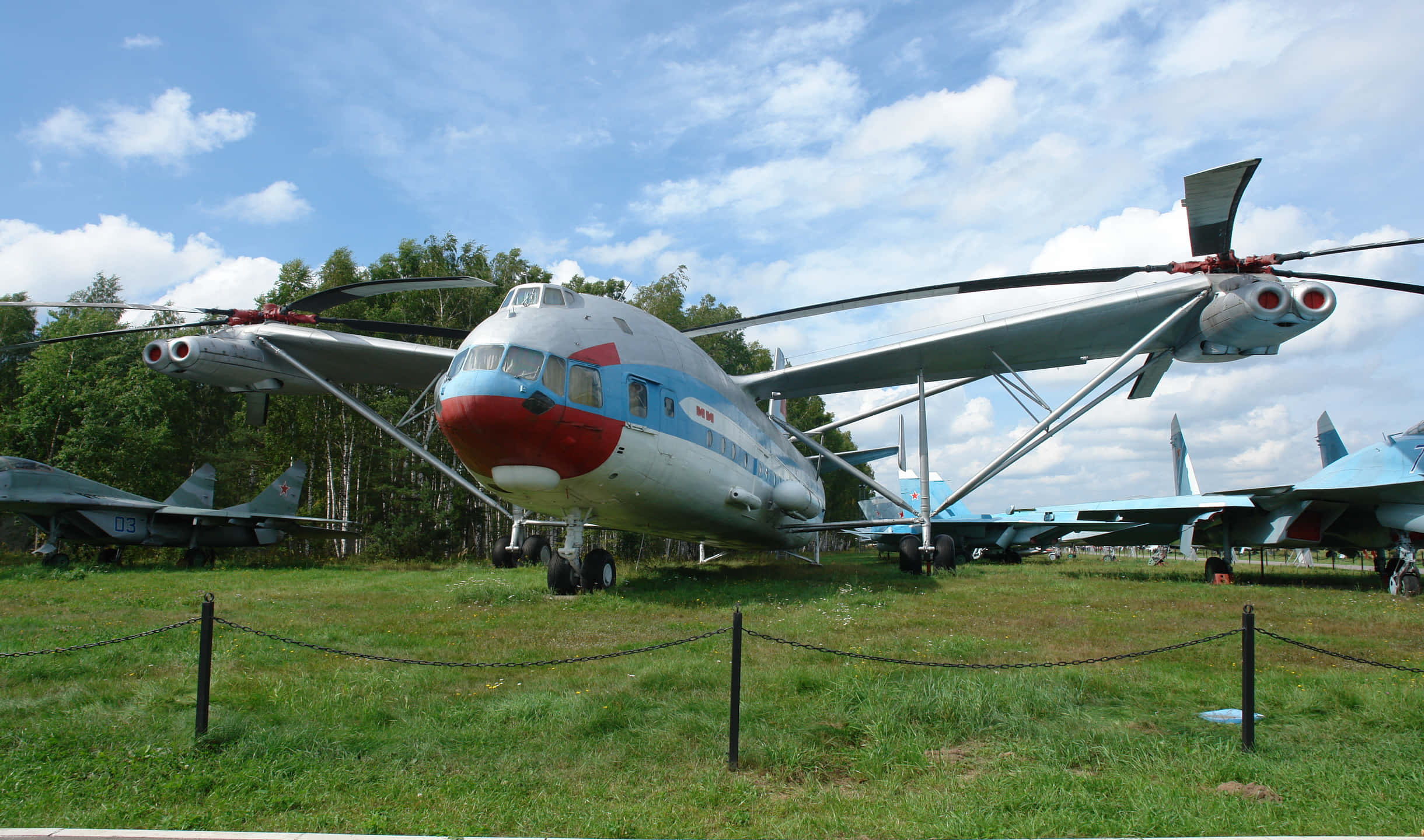 2440x1440 Helicopters 桌布 壁紙 — 一架大型蘇聯時代的直升機，紅色機頭和藍色條紋，停在草地上，周圍有其他軍用飛機，天空部分多雲。