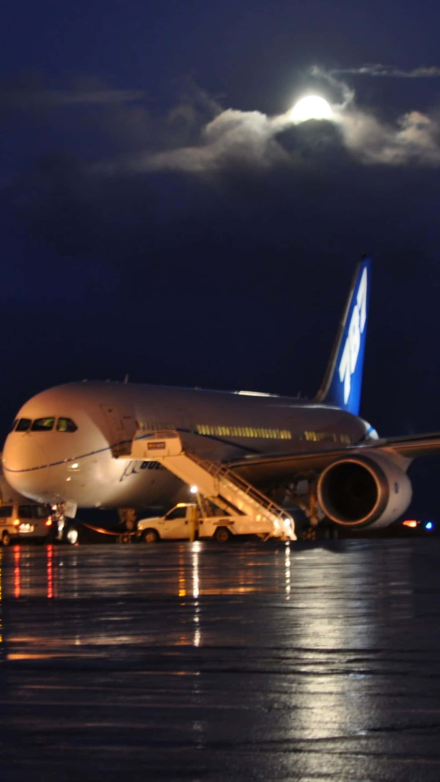 Plane 桌布 壁紙 — 一架大型飛機在夜晚停放在濕滑的停機坪上，明亮的月亮和雲朵下，地面燈光反射在路面上。