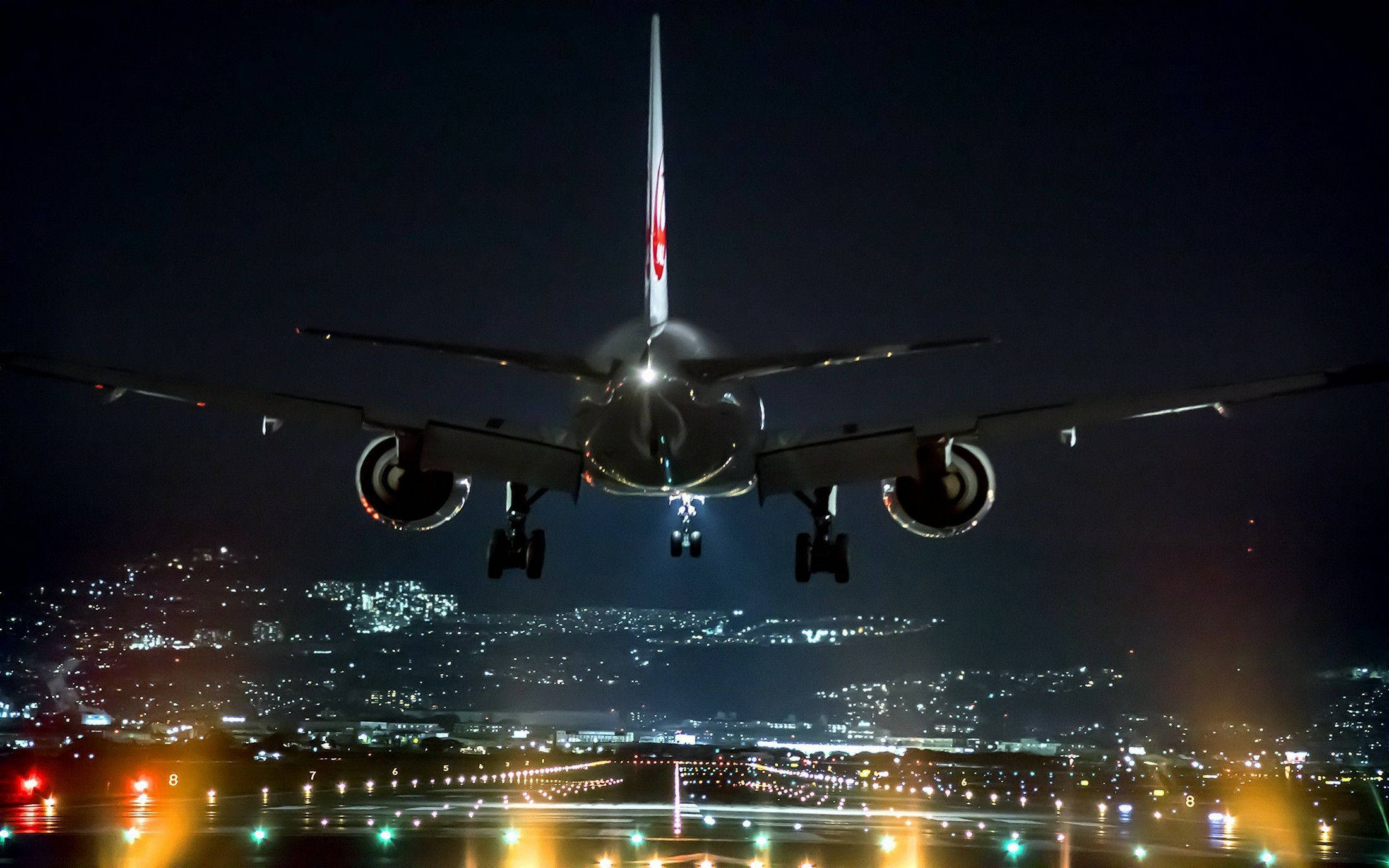 飛機 桌布 壁紙 — 一架大型飛機在夜間下降，下方濕滑的跑道上城市燈光閃爍。