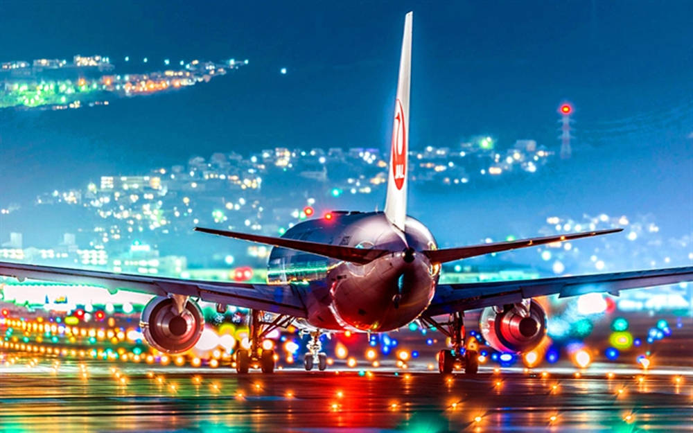 飛機 桌布 壁紙 — 一架大型飛機在夜間停在潮濕的跑道上，背景是充滿活力的城市景觀在發光。