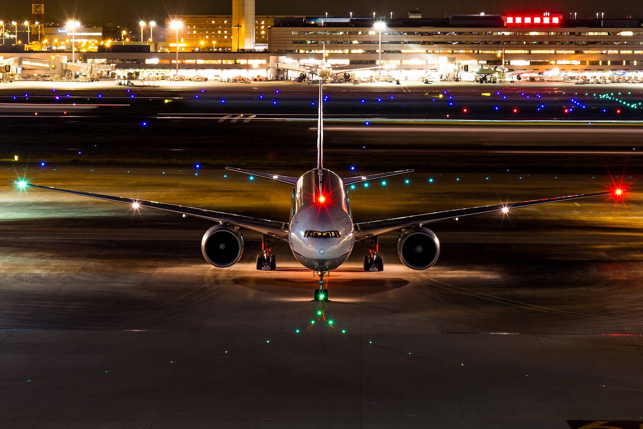 飛機 桌布 壁紙 — 一架大型飛機在夜間停在跑道上，被發光的燈光照亮，背景是燈火通明的機場航廈。