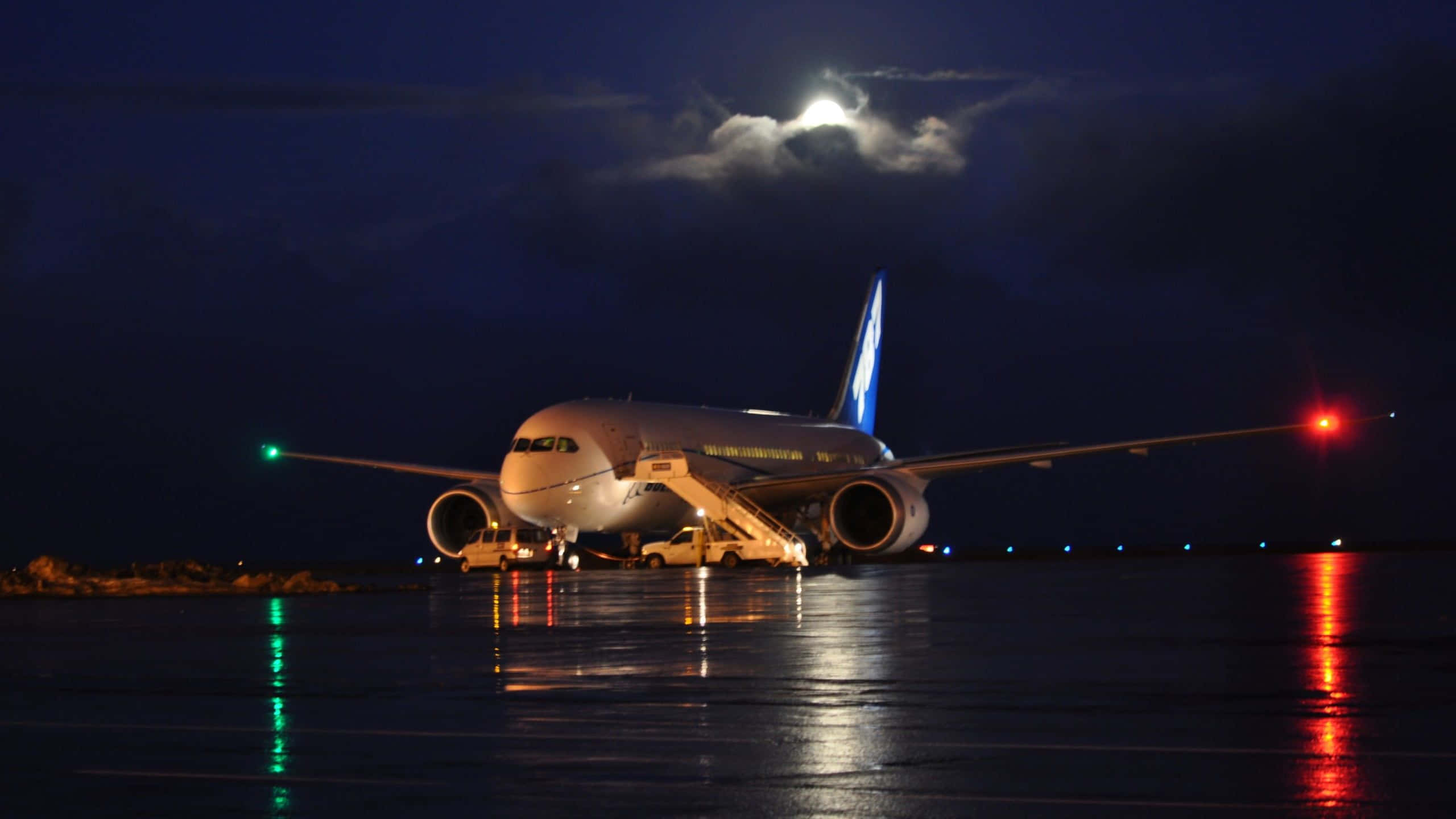 飛機 桌布 壁紙 — 一架大型飛機在夜間停泊在濕滑的停機坪上，明亮的月亮和雲朵下，紅色和綠色的燈光反射在地面上。