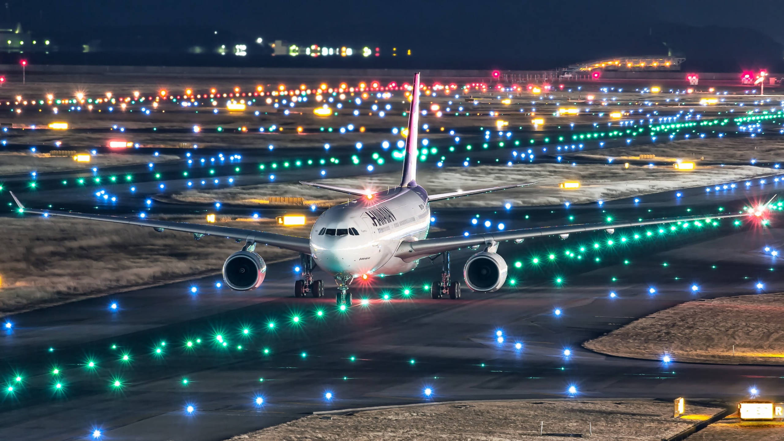 Runway 桌布 壁紙 — 一架大型飛機在夜間於一條被鮮豔綠色和藍色燈光照亮的跑道上滑行，遠處的機場建築物在背景中發光。