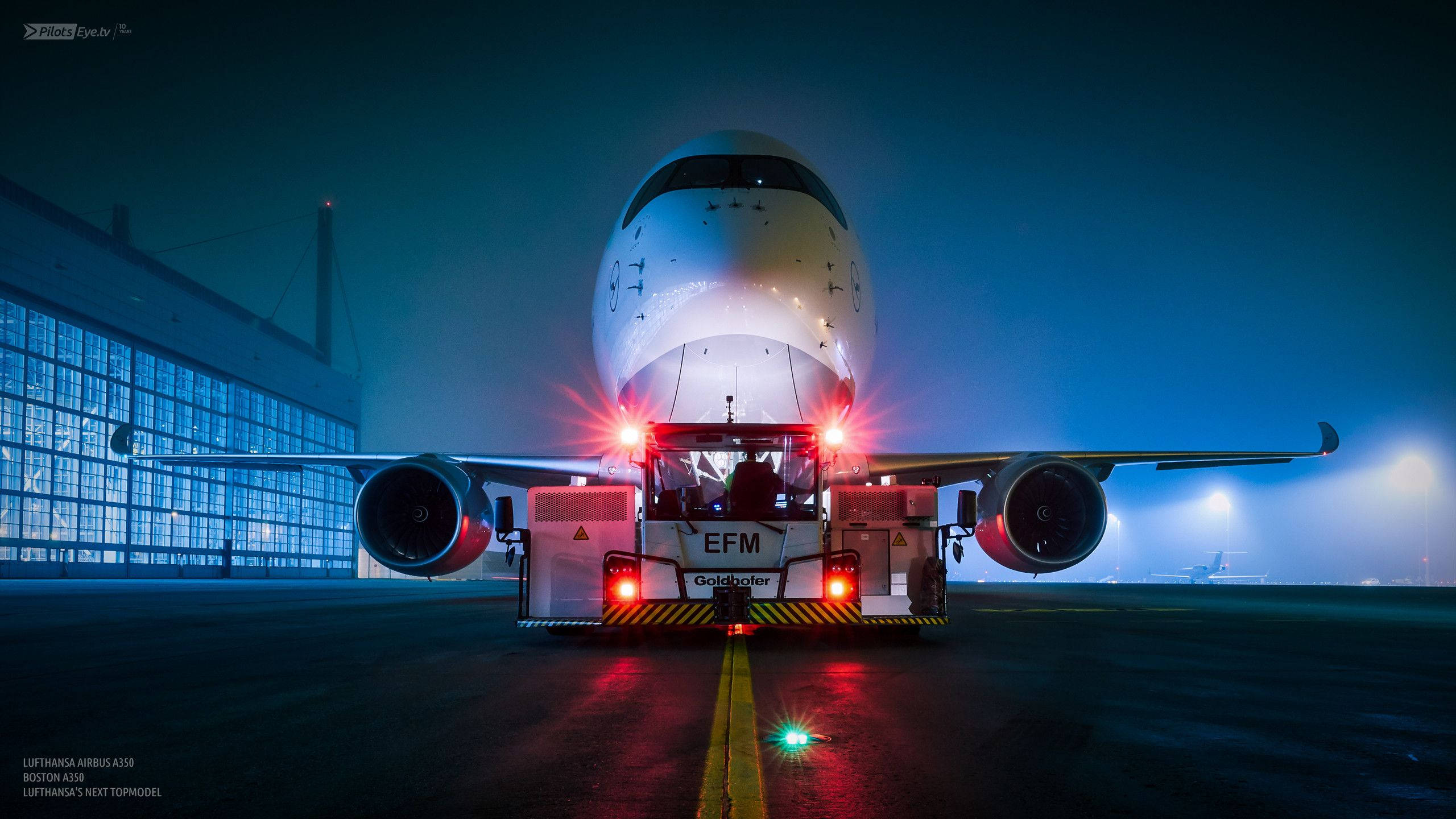 4K 飛機壁紙 桌布 壁紙 — 一架大型飛機在夜間於有霧的停機坪上被推後，背景有發光的紅燈和藍色調的機庫。