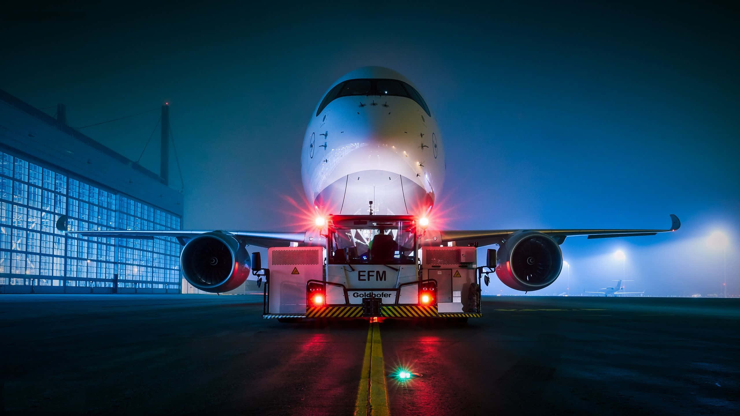 4K 飛機壁紙 桌布 壁紙 — 一架大型飛機在夜間被拖行在潮濕的停機坪上，背景有發光的紅燈和藍色機庫。