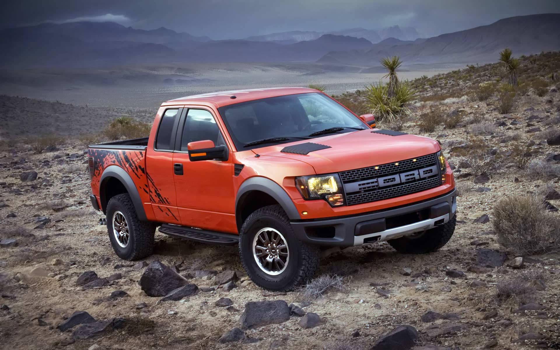 Trucks 桌布 壁紙 — 一架大膽的橙色Ford F-150 Raptor在陰沉多雲的天空下，堅固地矗立在岩石沙漠地形上。