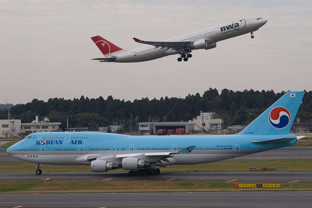 Airport 桌布 壁紙 — 一架大韓航空波音747在跑道上滑行，同時一架大韓航空空中巴士A330在頭頂起飛，背景是陰天。