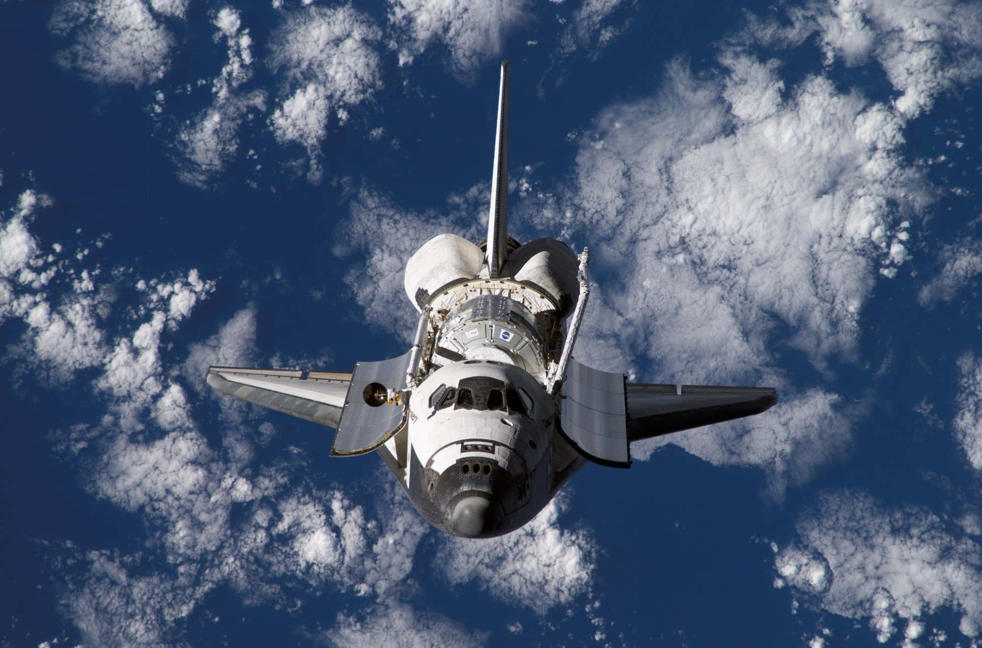 Space Mission 桌布 壁紙 — 一架太空梭在地球上空翱翔，蓬鬆的白雲在其機翼下飄動，襯托著深藍色的天空。