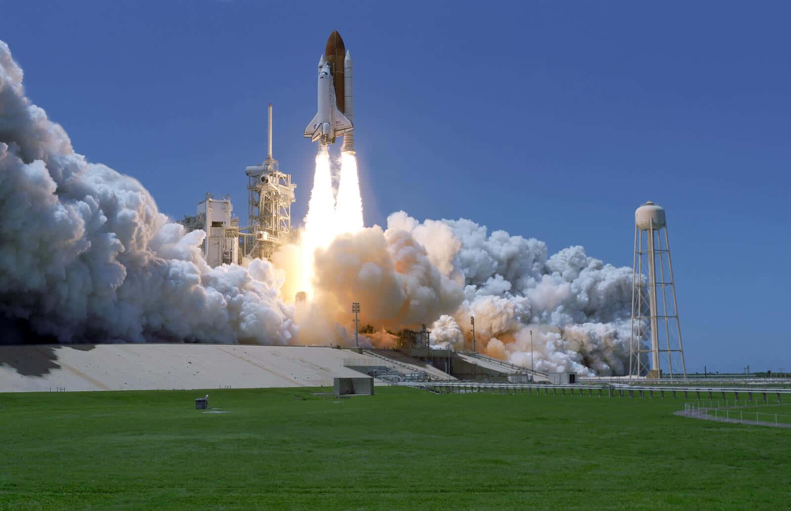 Space Mission 桌布 壁紙 — 一架太空梭在晴朗的藍天中發射，從長滿草地的發射台上升時噴出濃厚的白色煙霧。